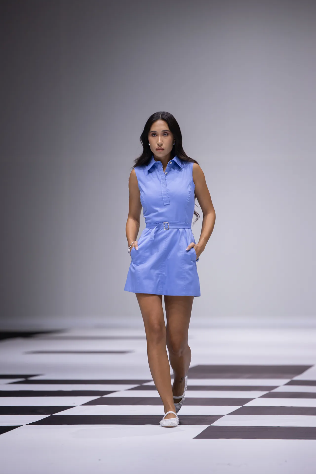 Woman in a light blue sleeveless belted dress walking down a black-and-white checkered runway.