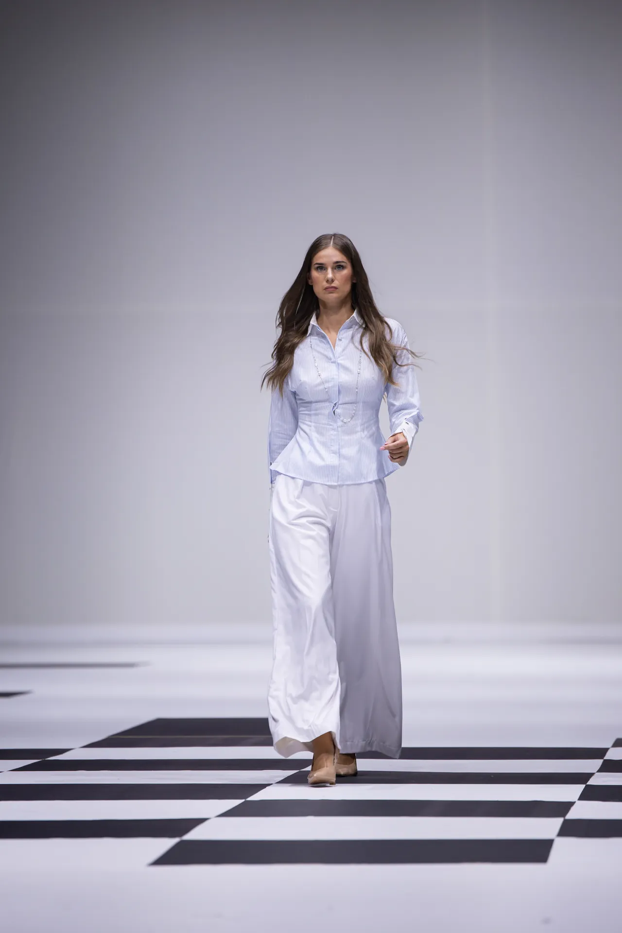 Female model walking a fashion runway on a black-and-white checkerboard floor, wearing a light blue blouse and white wide-leg pants.