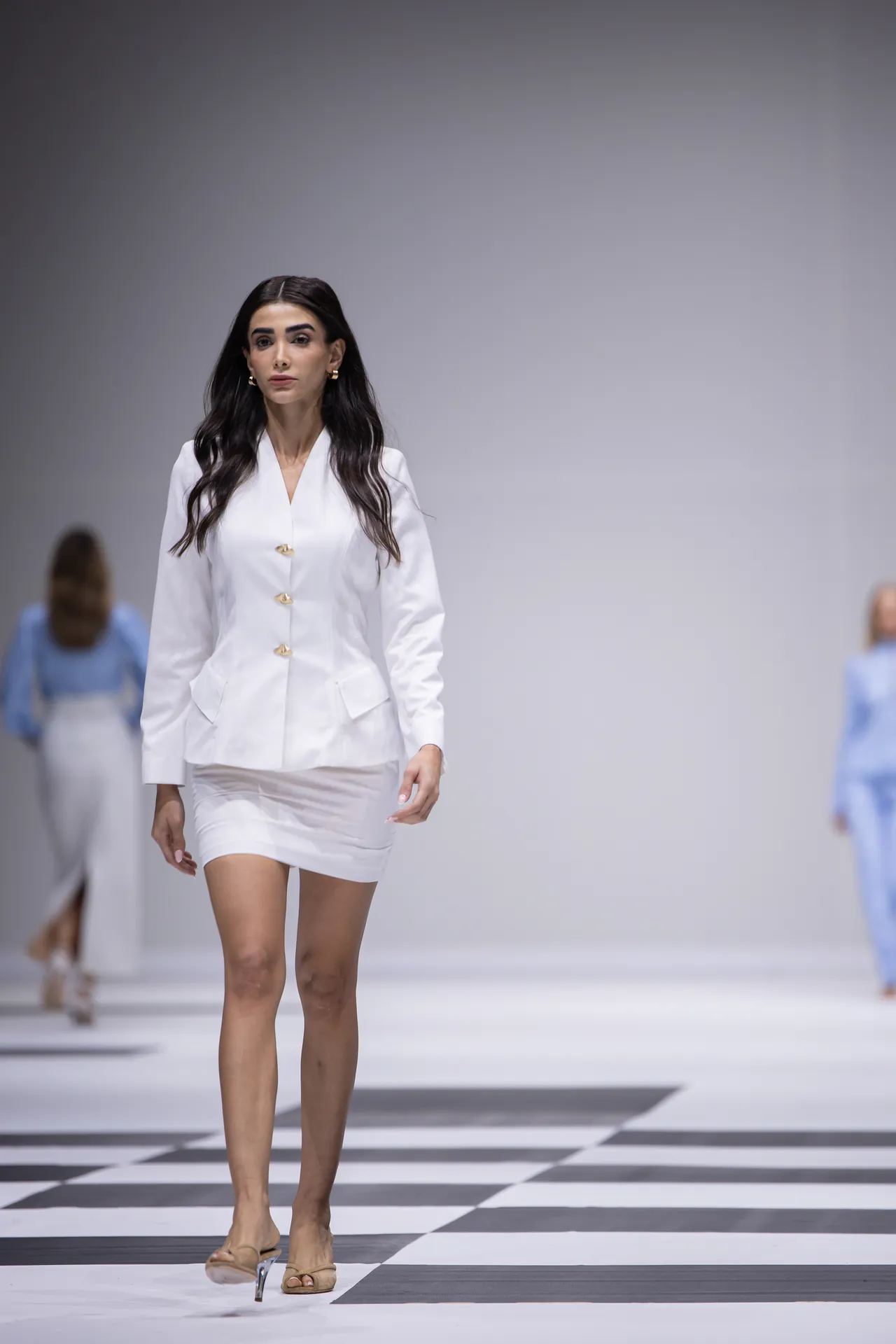 Model walks a white blazer and matching mini skirt on a runway with a black-and-white checkered floor, neutral heels, and dark hair.