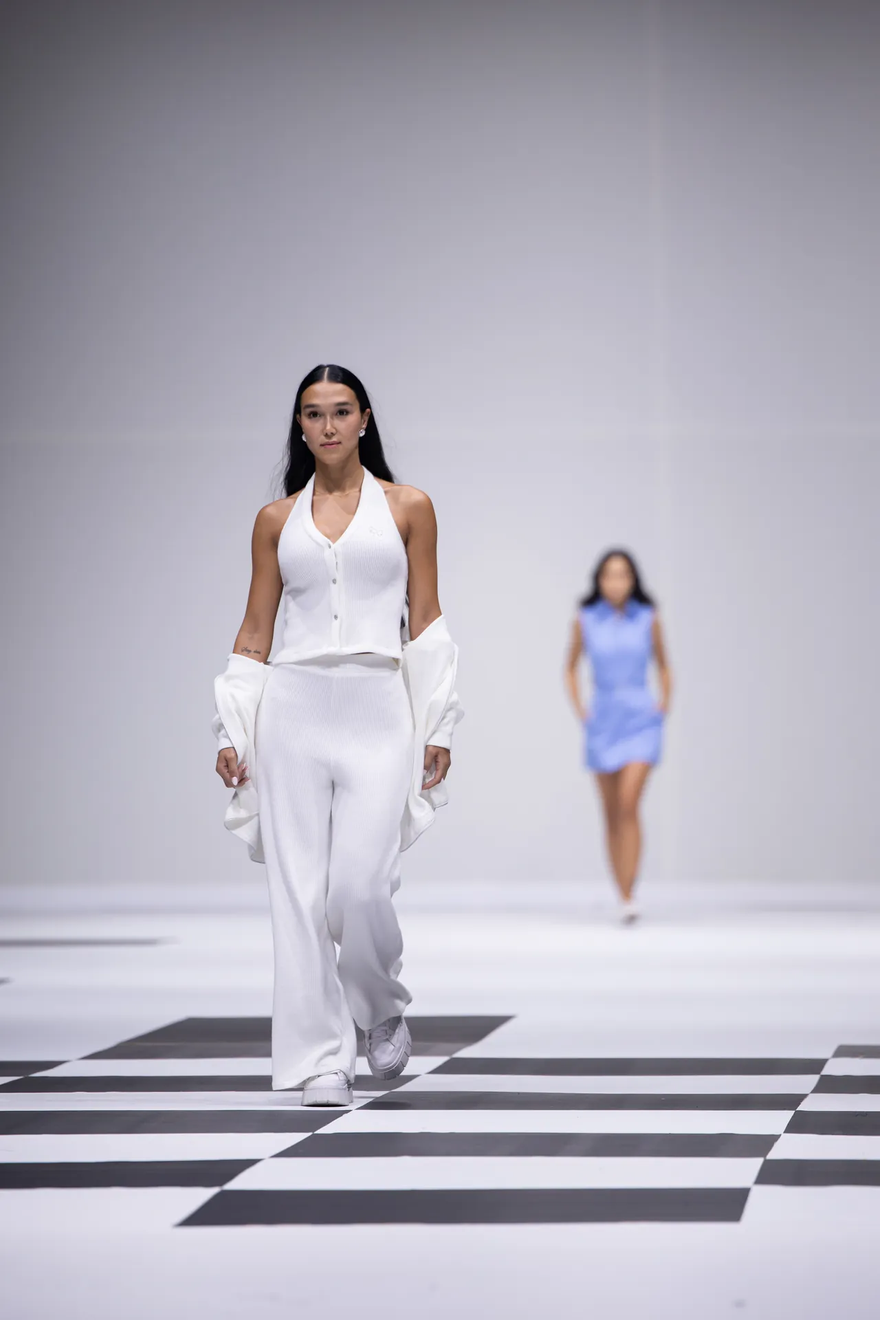 Female model in white halter-top and wide-leg pants walks a black-and-white checkered runway, backstage lighting neutral.