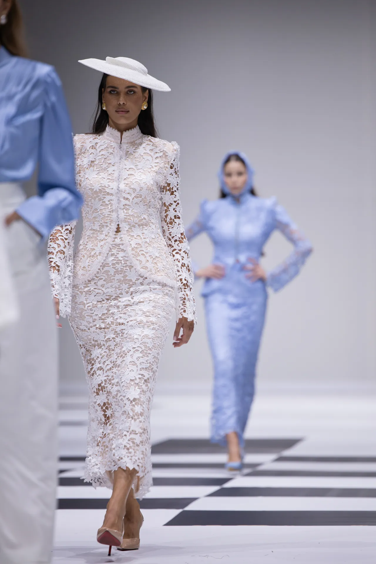 Model walks a runway in a white lace full-length dress and wide-brim white hat, nude heels, against a black-and-white checkered floor. Behind her, another model in blue dress watches in the distance.