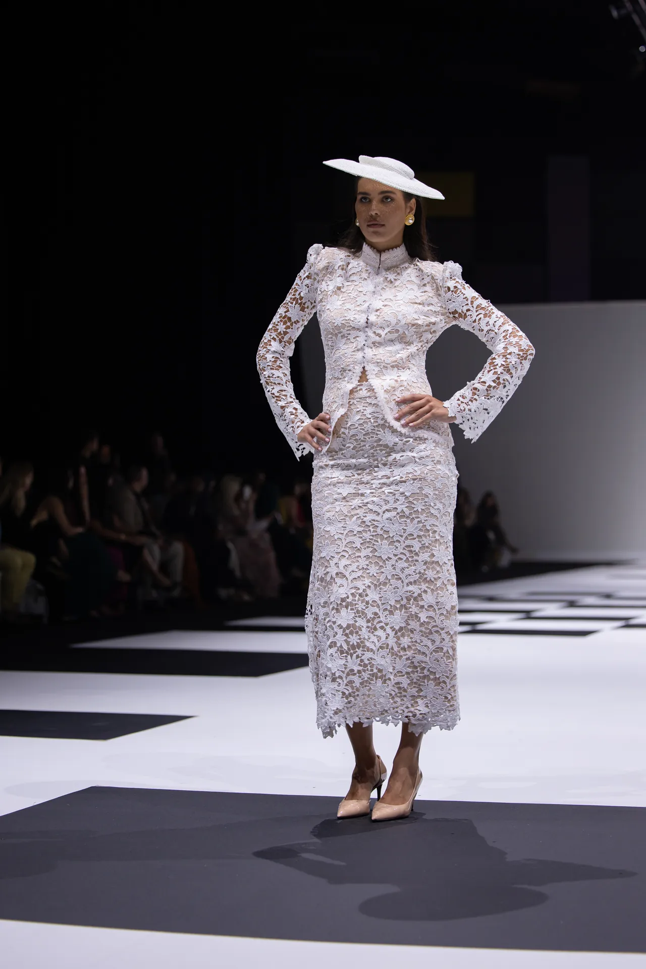Model on a fashion runway wearing a white lace long-sleeve midi dress with a wide-brim white hat and beige heels, posing with hands on hips.