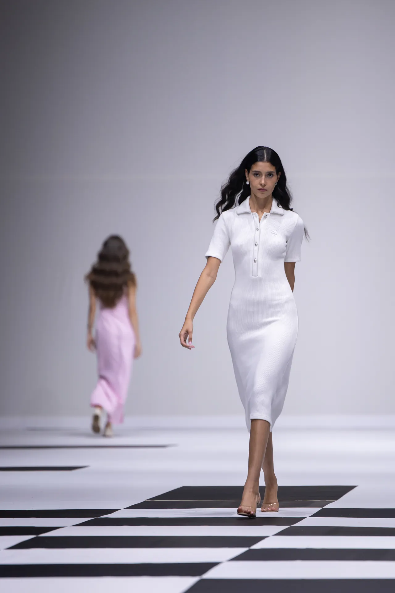 Model walks down a minimalist runway wearing a white short-sleeve knit midi dress; black-and-white checkered floor visible underfoot.