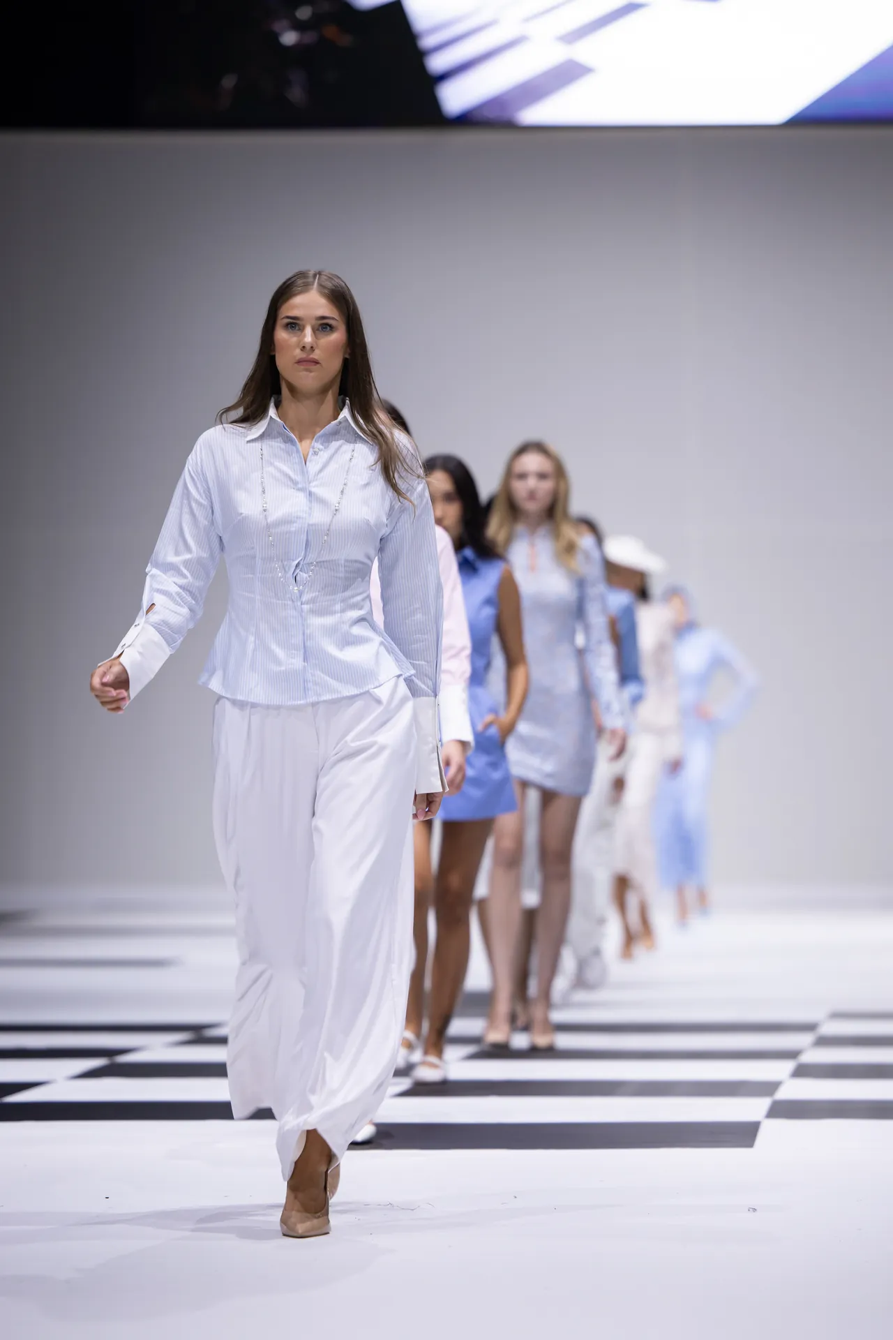Model walks the runway in a pale blue striped shirt and white wide-leg pants, with more models in blue and white outfits behind on a black-and-white checkered floor.