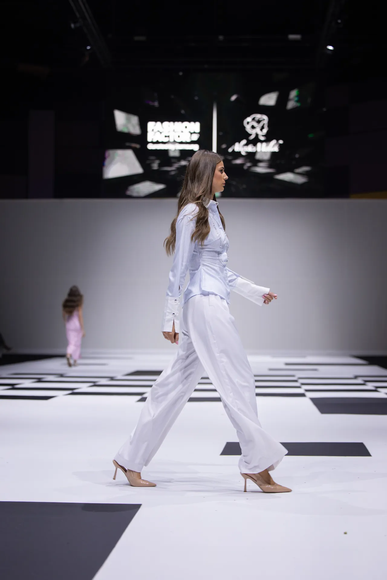 Female model in a white pantsuit walking on a black-and-white checker runway.