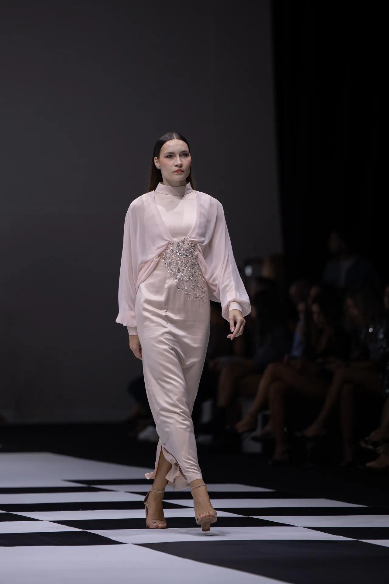 Model walks a pale pink gown with a jeweled belt down a black-and-white checkerboard runway.