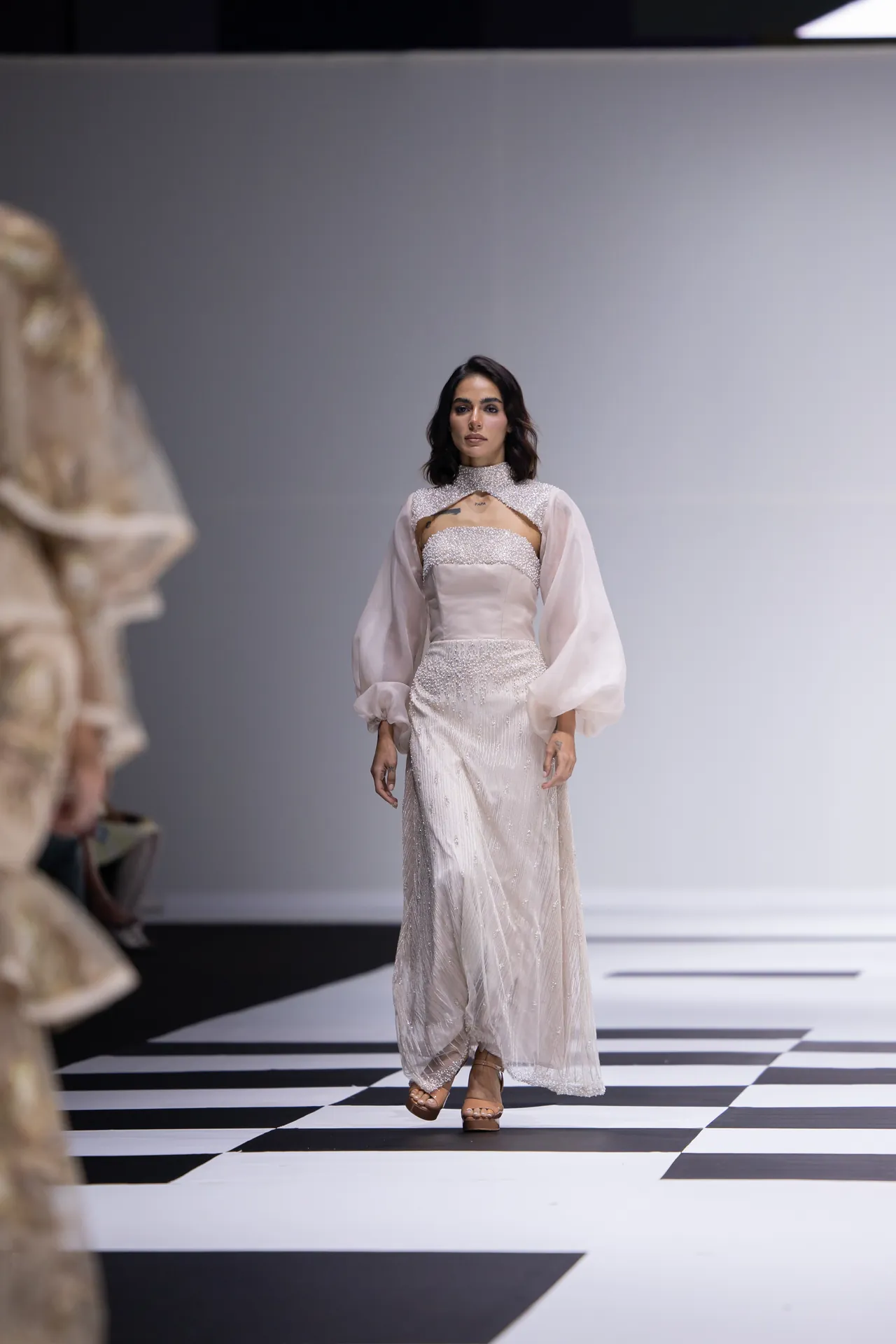 Model walking the runway in an ivory beaded gown with sheer puff sleeves.