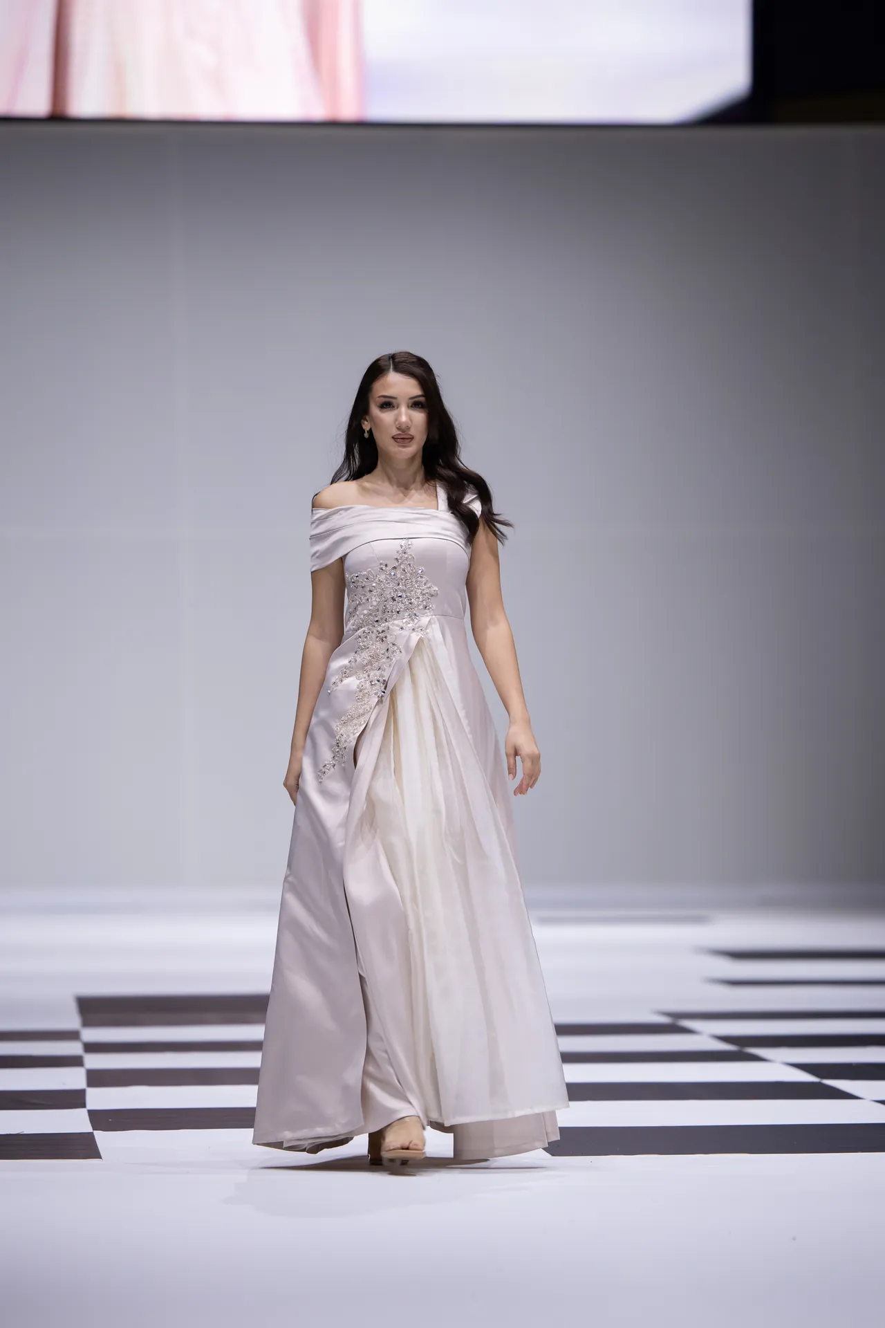 Model wearing a light off-shoulder gray evening gown with embroidery walking on a fashion runway with a black-and-white checkered floor.