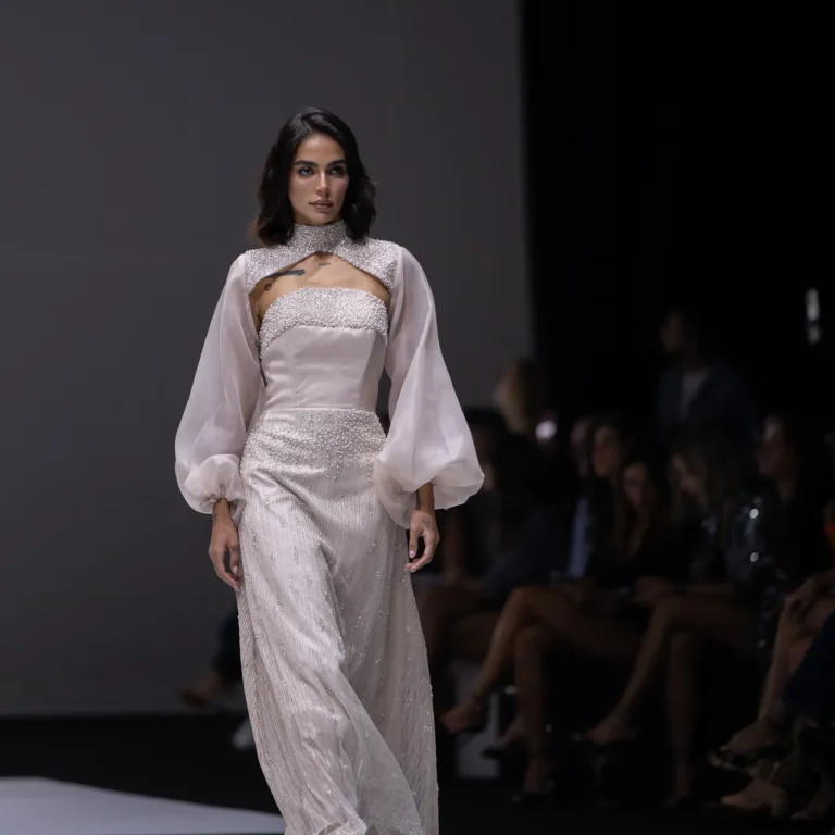 Model walks a white beaded gown with sheer puff sleeves on a black-and-white checkered runway.