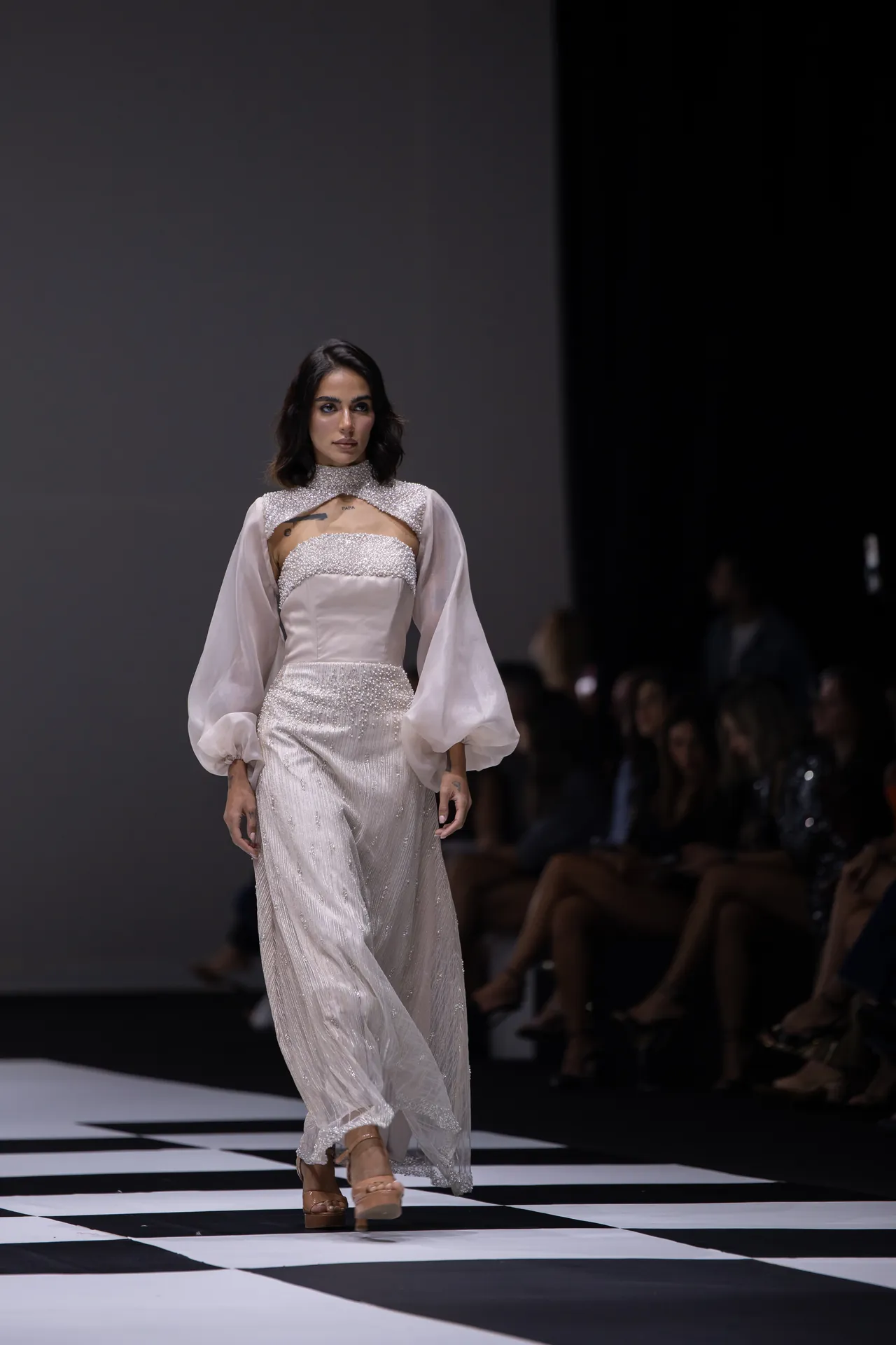 Model walks a white beaded gown with sheer puff sleeves on a black-and-white checkered runway.