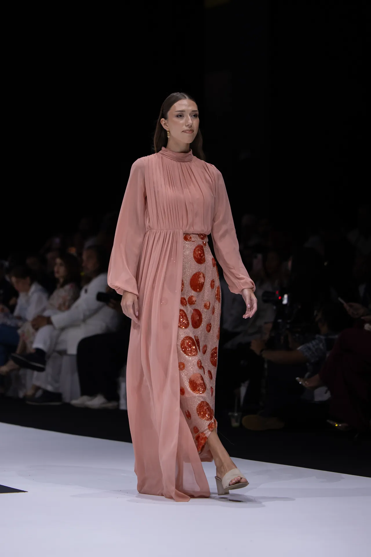 Model on a fashion show runway wearing a pink long-sleeve pleated gown with a sheer cape and red circular sequins, audience in dim background.