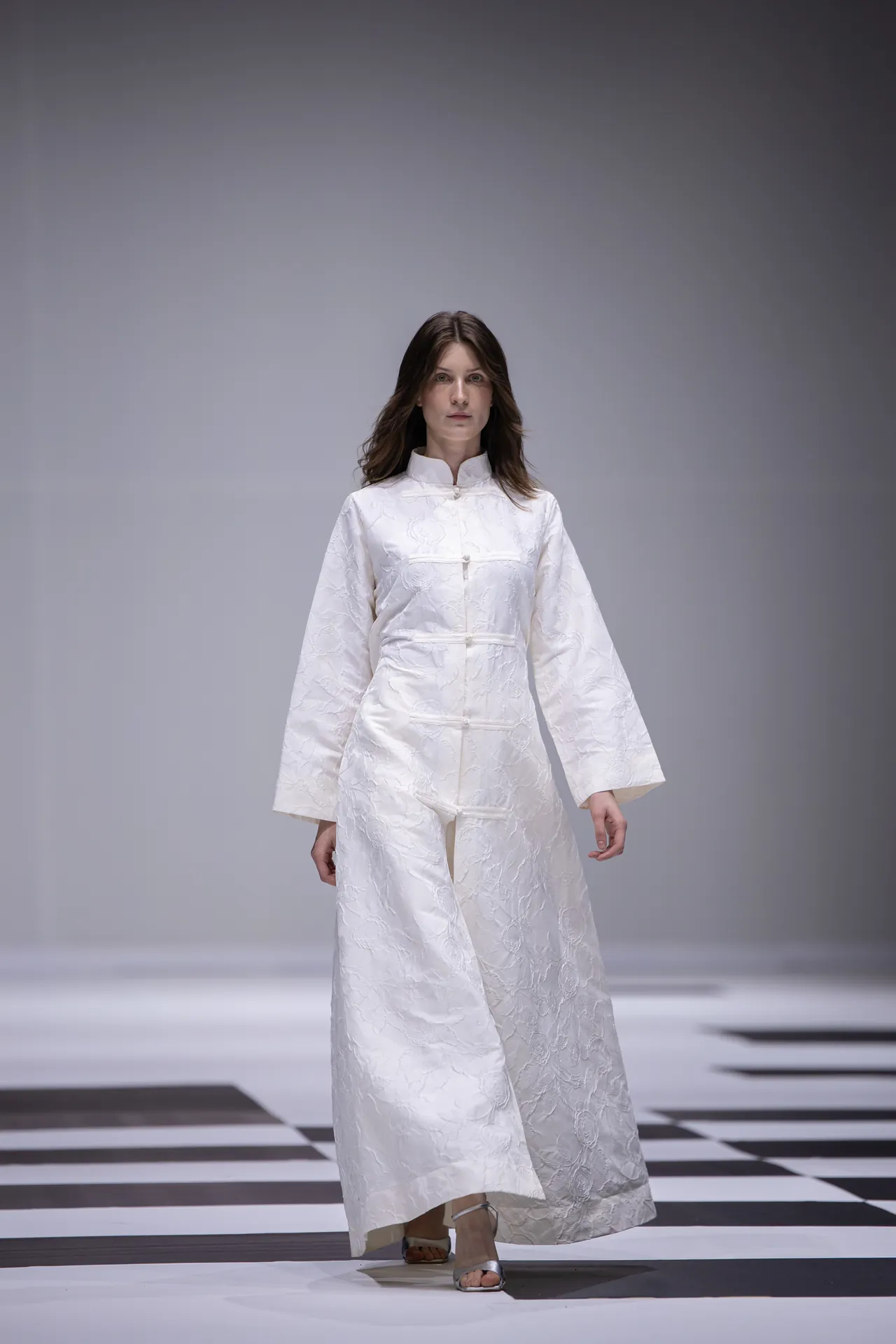 Model walking a fashion runway in a long white textured robe with a high collar and wide sleeves on a black-and-white checkered floor.