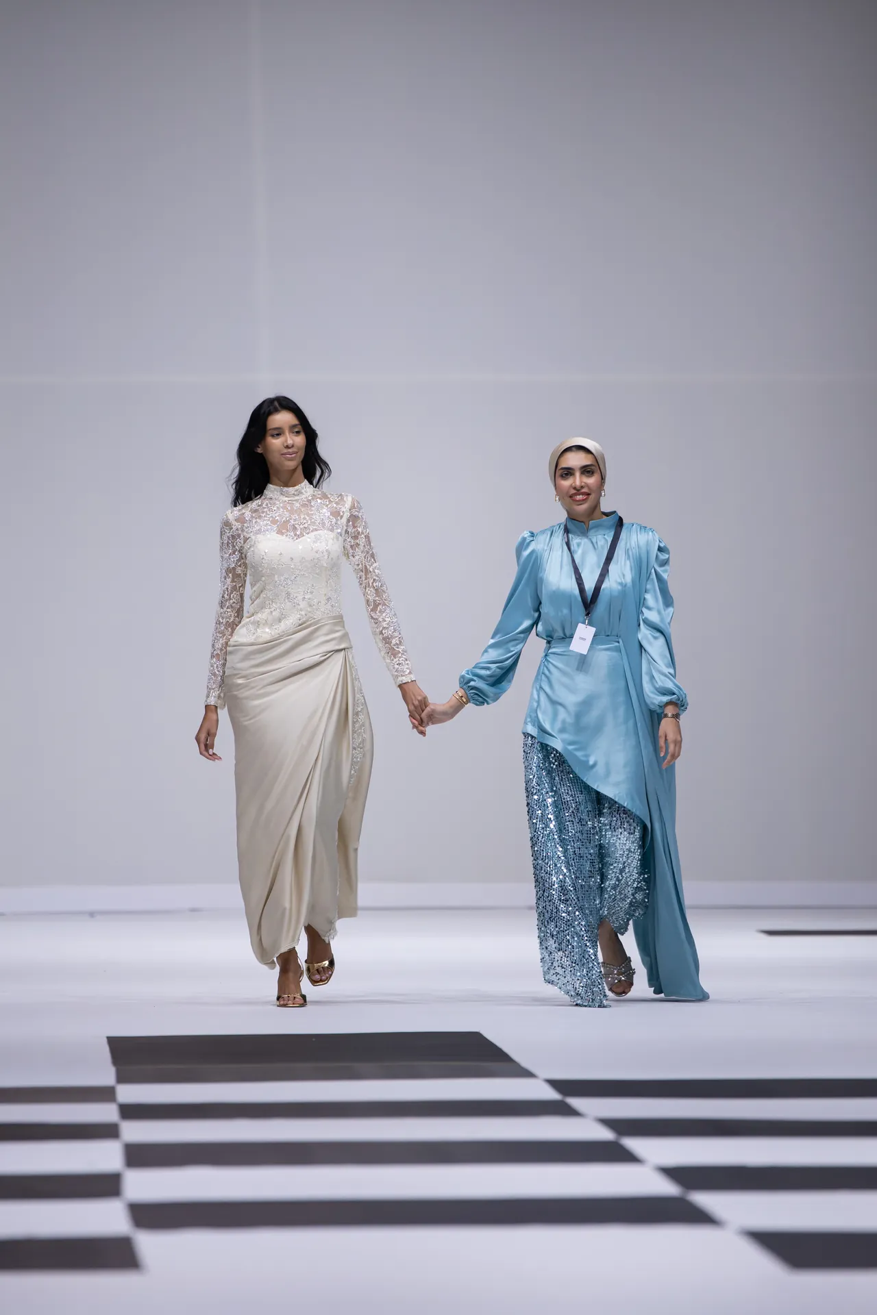 Two women walk hand in hand down a fashion runway; one wears a cream lace gown, the other a blue satin dress with a hijab and badge.