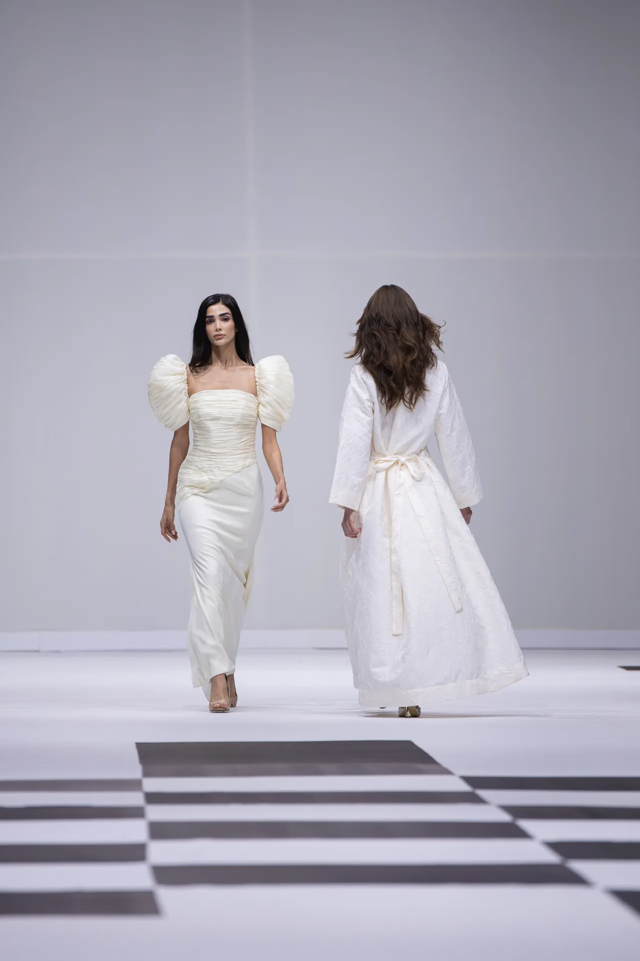 Two models walk a white fashion runway in elegant white gowns; one faces the camera with a structured puff-sleeve dress, the other walks away in a long textured gown.