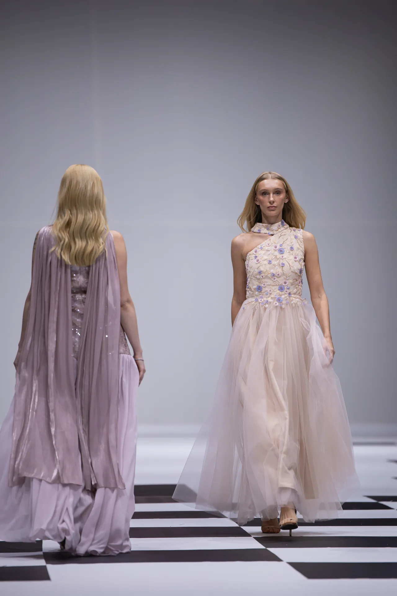 Runway scene with two models in pastel gowns: one facing camera in a pale pink floral-embellished dress, the other seen from behind in a lavender cape dress on a black-and-white checkered floor.