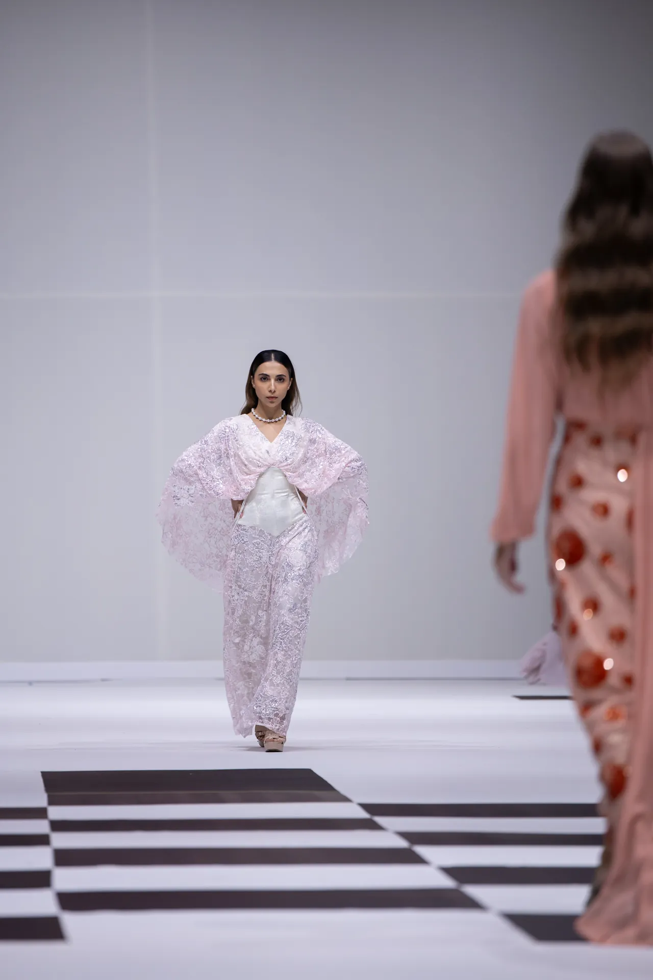 Model walks the fashion runway in a pale pink lace jumpsuit with dramatic cape sleeves.