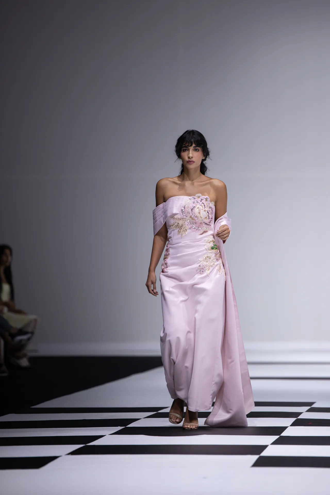 Model walking a pale pink off-shoulder gown with floral embroidery on a black-and-white checkered runway.