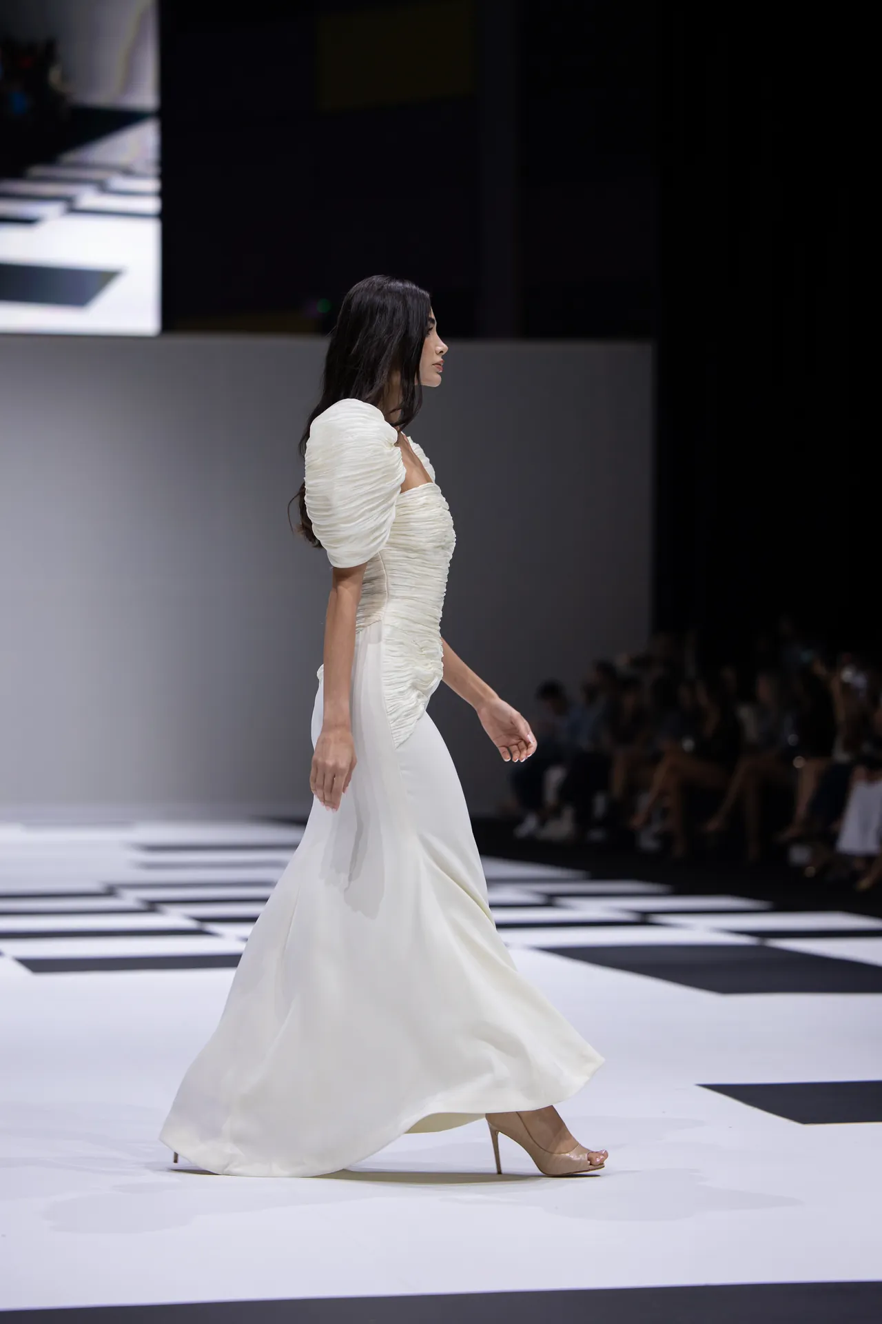 Model walks a white wedding-style gown with ruched bodice and puff sleeves on a fashion runway.