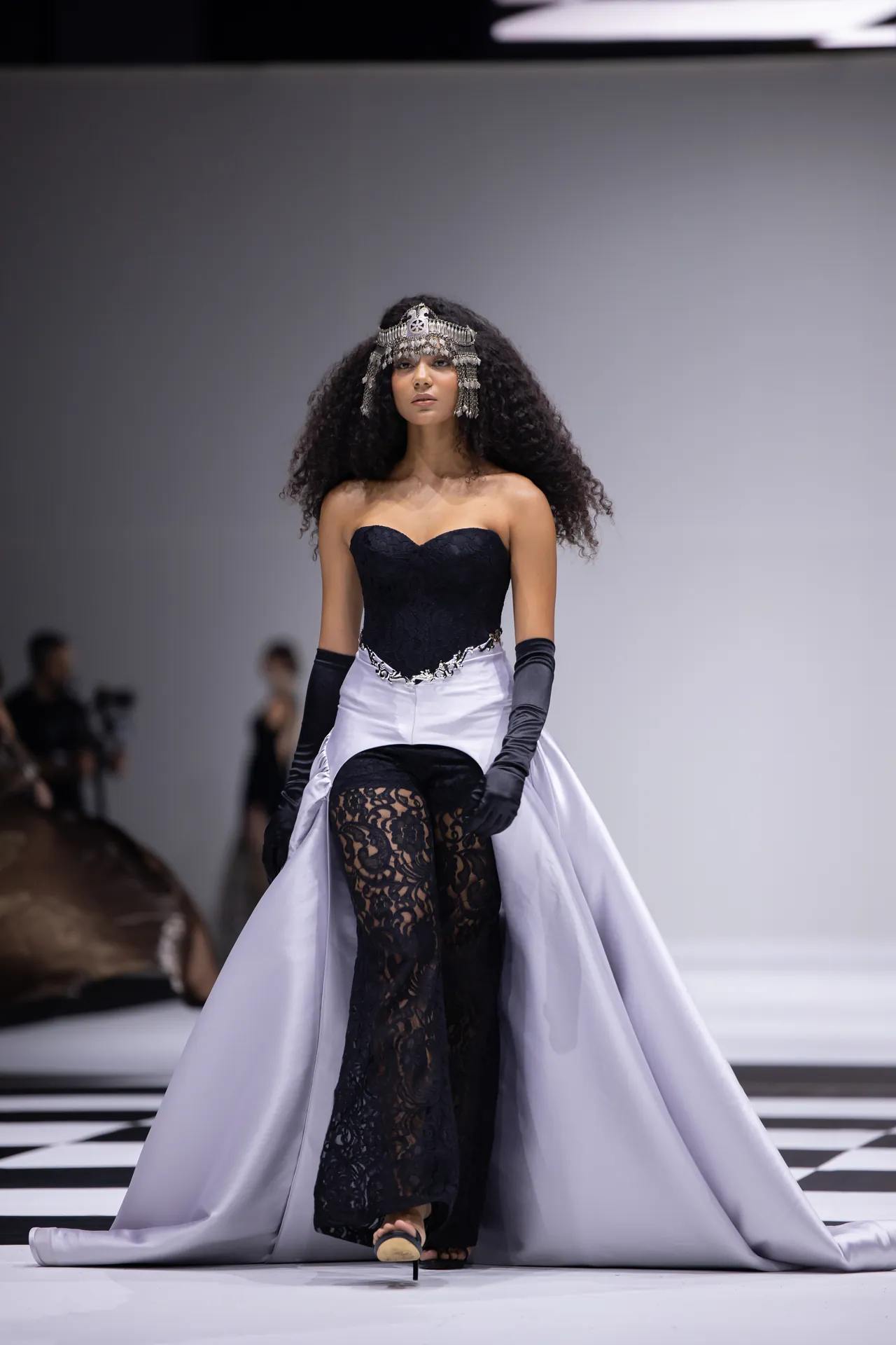 Model walking a runway in a strapless black bodice, pale lavender train skirt, black lace tights, gloves, and an ornate silver headpiece.
