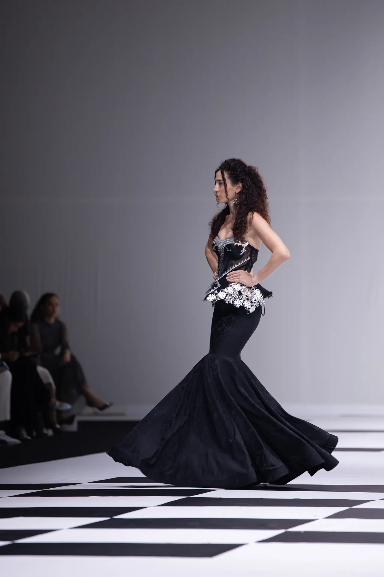 Model wearing a black mermaid gown with white floral embroidery walks the runway on a black-and-white checkered floor.