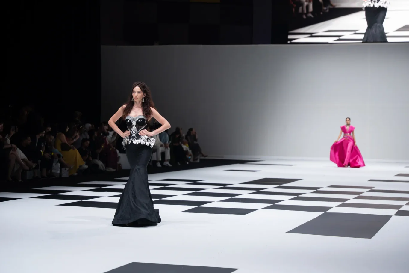 Model in a black, ornate mermaid gown walks a white and black checkered runway; audience seated along the left. (fashion show)