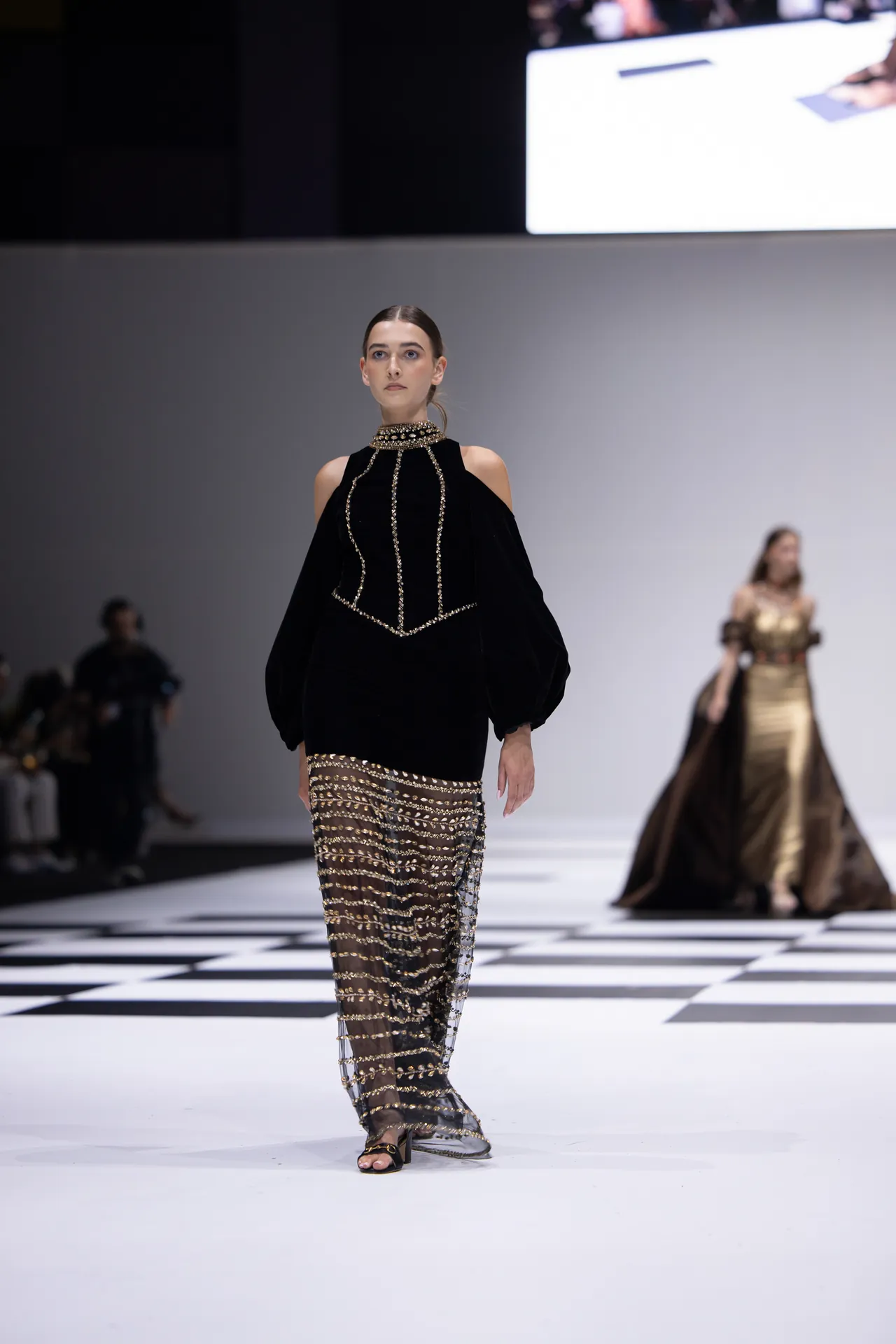 Model on a runway wearing a black velvet top with gold beaded trim and sheer gold-sequined pants, checkerboard floor underfoot.
