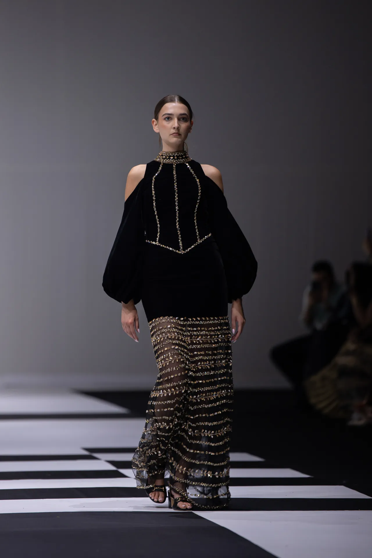 Model walking the runway in a black cold-shoulder gown with gold beading and a sheer, sequined skirt.