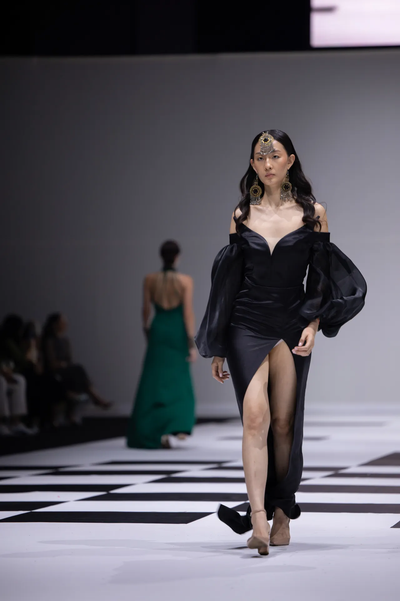 Model on a fashion runway in a black off-the-shoulder gown with dramatic sheer puff sleeves and large gold jewelry, walking forward on a checkered catwalk.