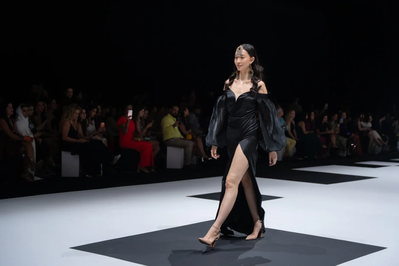 Model walks the runway in a black off-shoulder gown with dramatic sheer sleeves and gold headpiece, as an audience watches from the left.