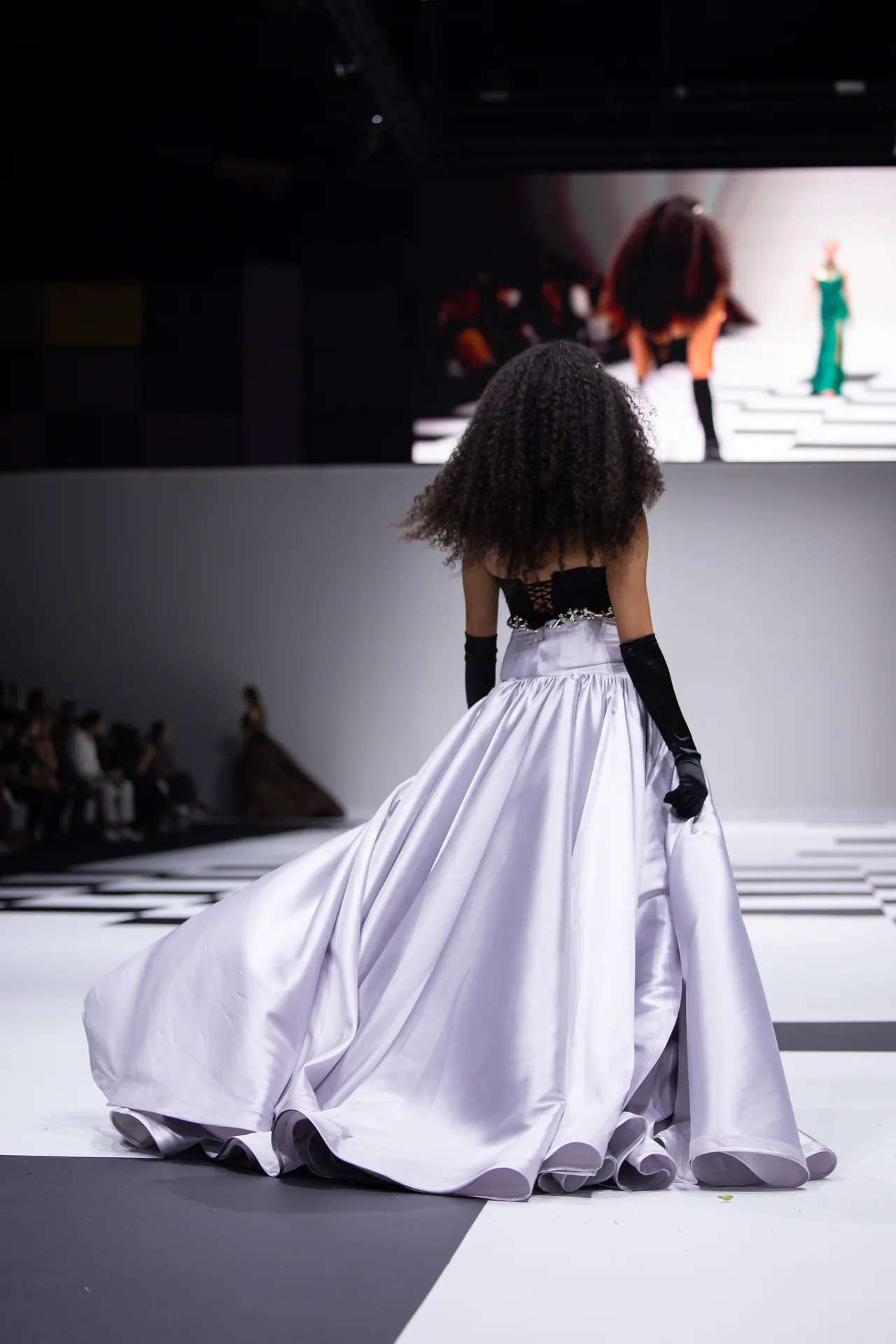 Model on a runway shown from behind, wearing a dramatic silver ballgown with black gloves, spotlight on the flowing skirt.