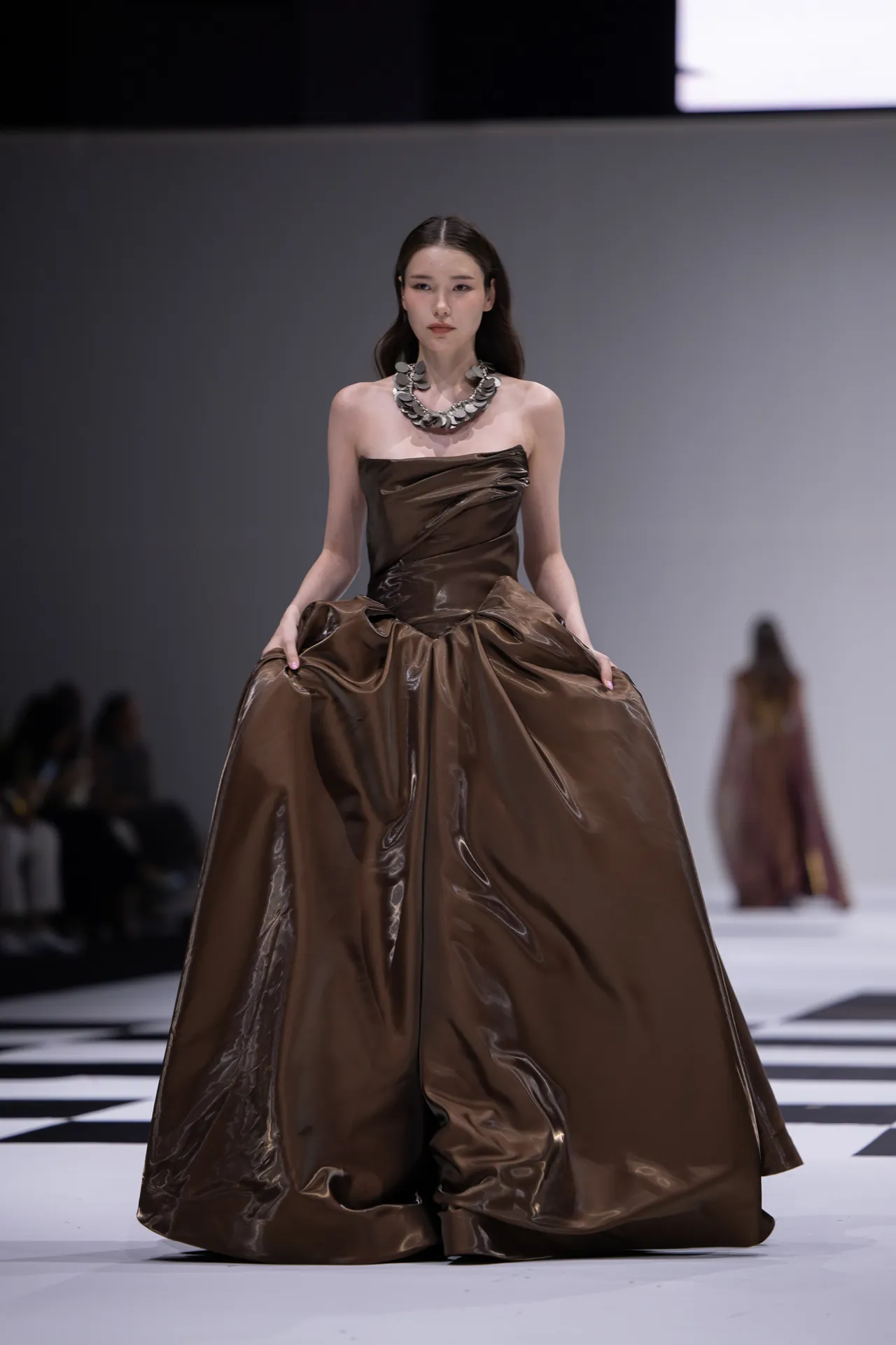 Model walks the runway in a glossy brown strapless ball gown with a bold silver necklace.