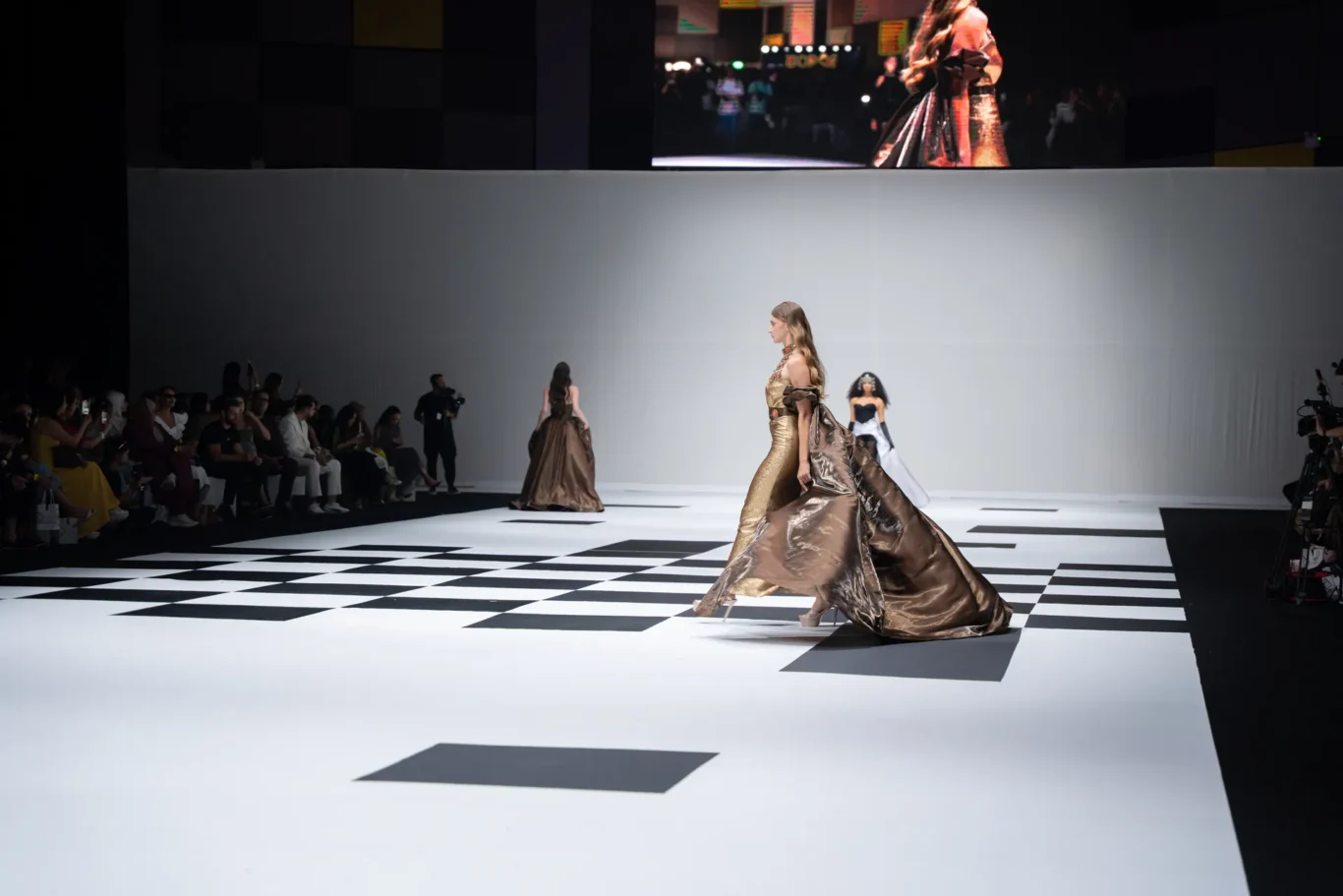 Model in a metallic gold gown with a long flowing train walks a black-and-white checkered runway; seated audience watches on the left, large screen above.