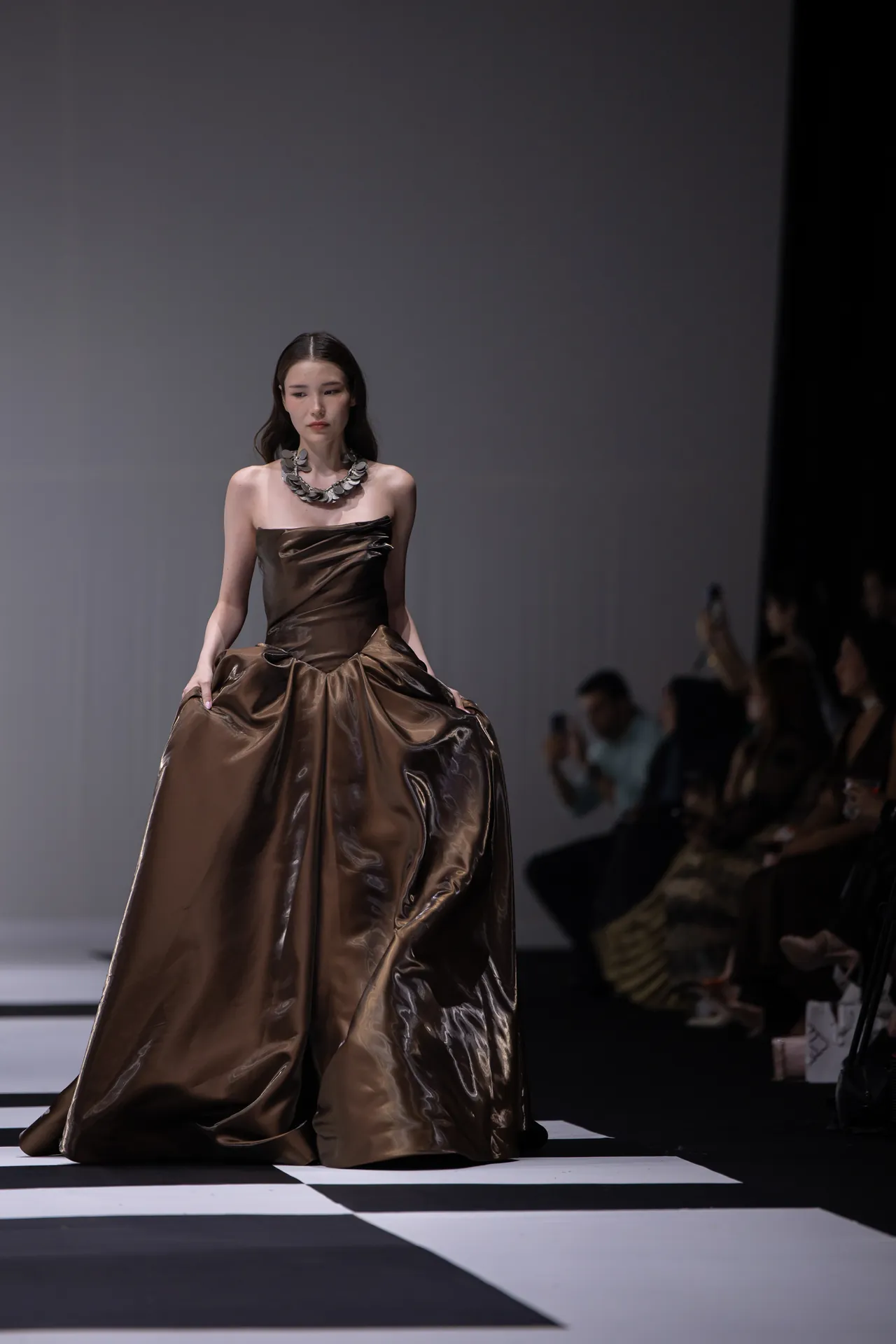 Model walks a bronze-brown metallic gown down the runway, wearing a chunky silver necklace.