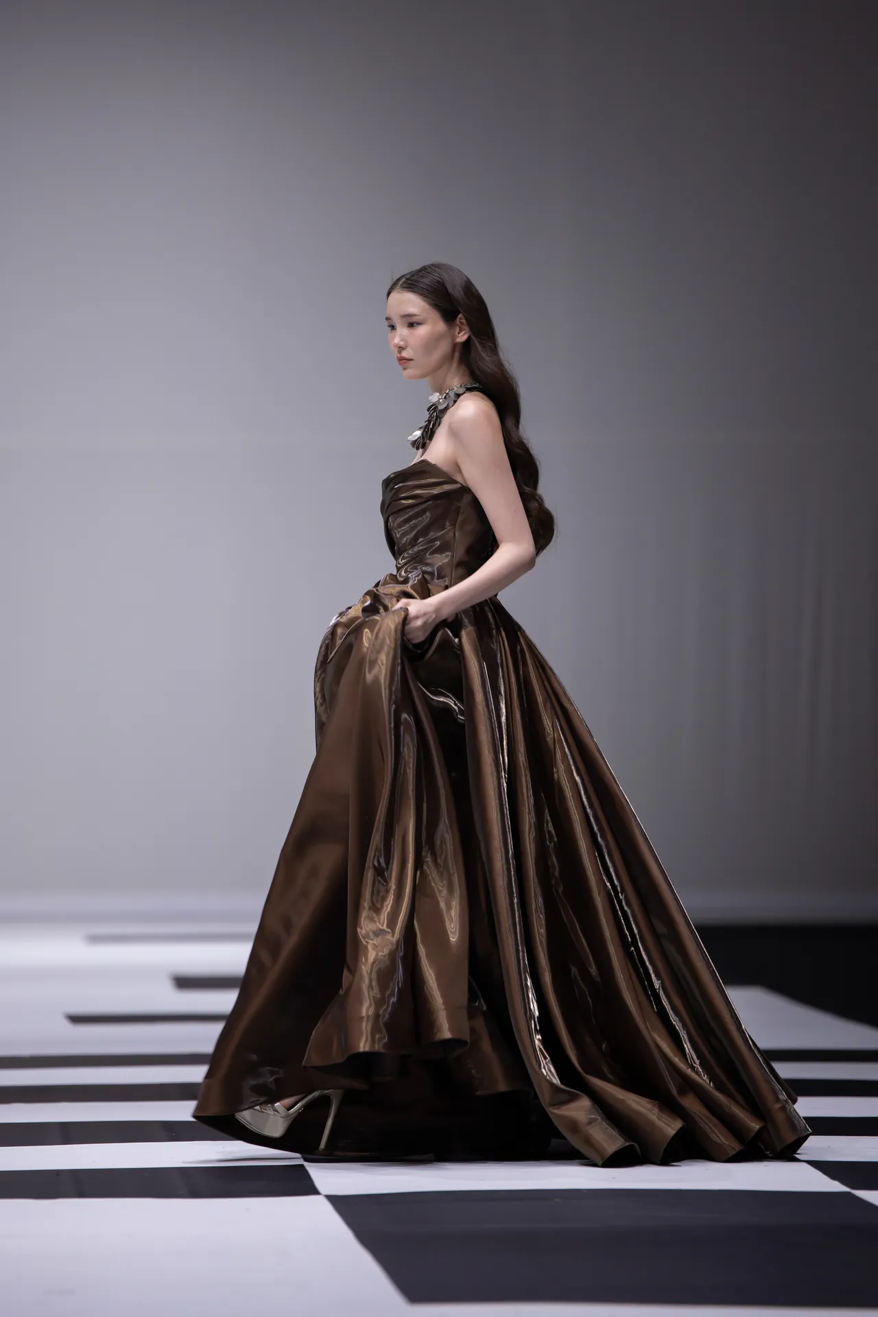 Model in a glossy bronze-brown strapless ball gown walking the runway, holding the dress to show its volume.