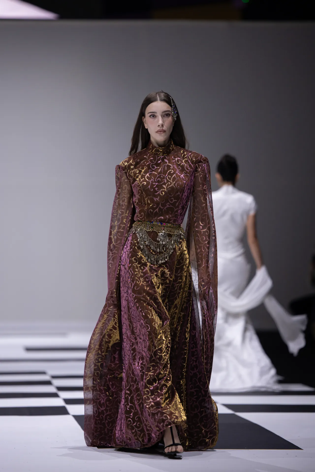 Model walks the runway in a sheer burgundy and gold patterned gown with an ornate belt accessorizing the waist.
