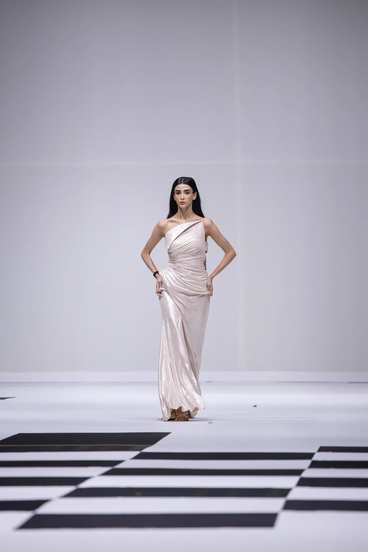 Model walks a runway in a one-shoulder, shimmery champagne gown, posing with hands on hips against a white backdrop.