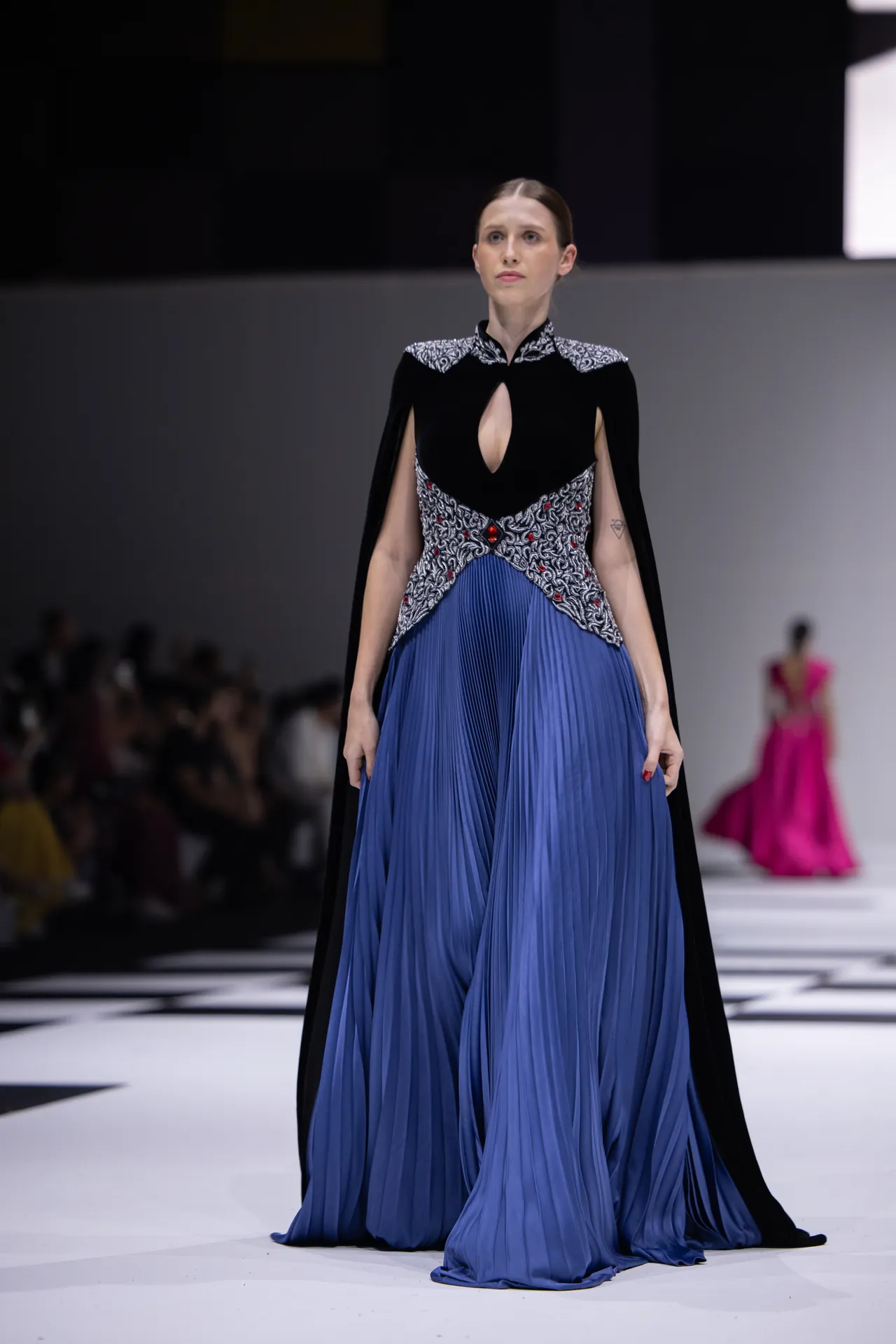 Model on a runway wearing a blue pleated gown with a black patterned bodice and cape.