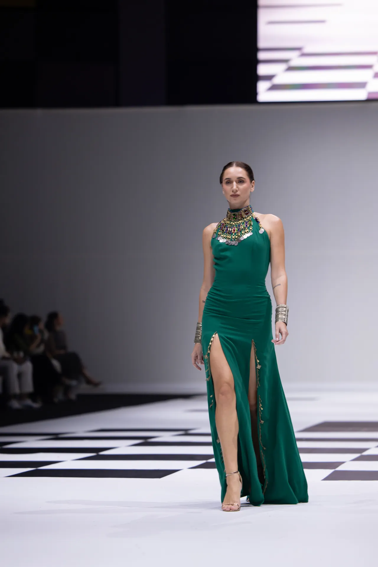 Runway model in an emerald-green gown with high slits and a large jeweled necklace, walking the catwalk on a checkered floor.
