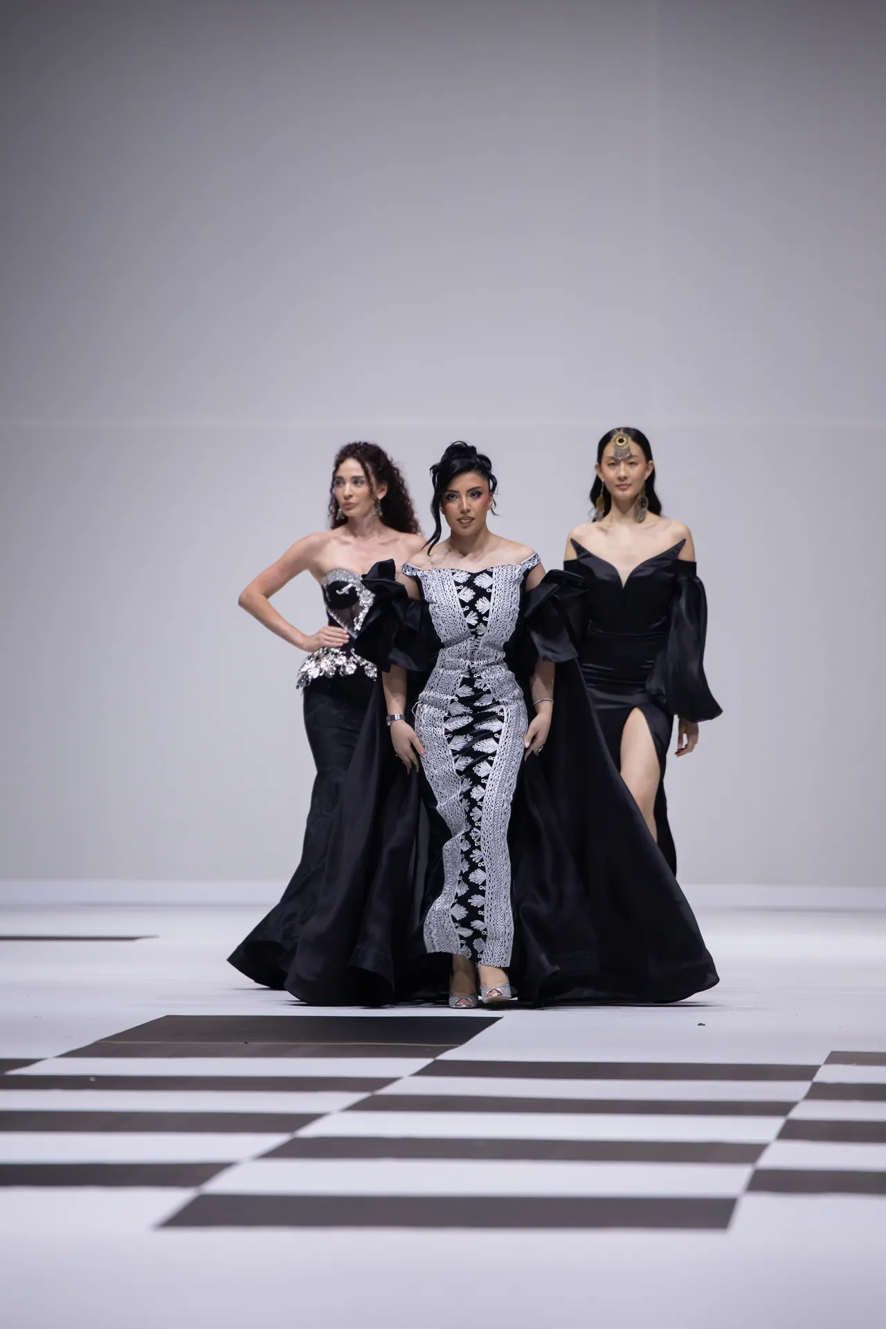 Three models walk a fashion show runway in black evening gowns; the center wears a silver-patterned off-shoulder dress, while the others wear sleek black gowns with intricate details.