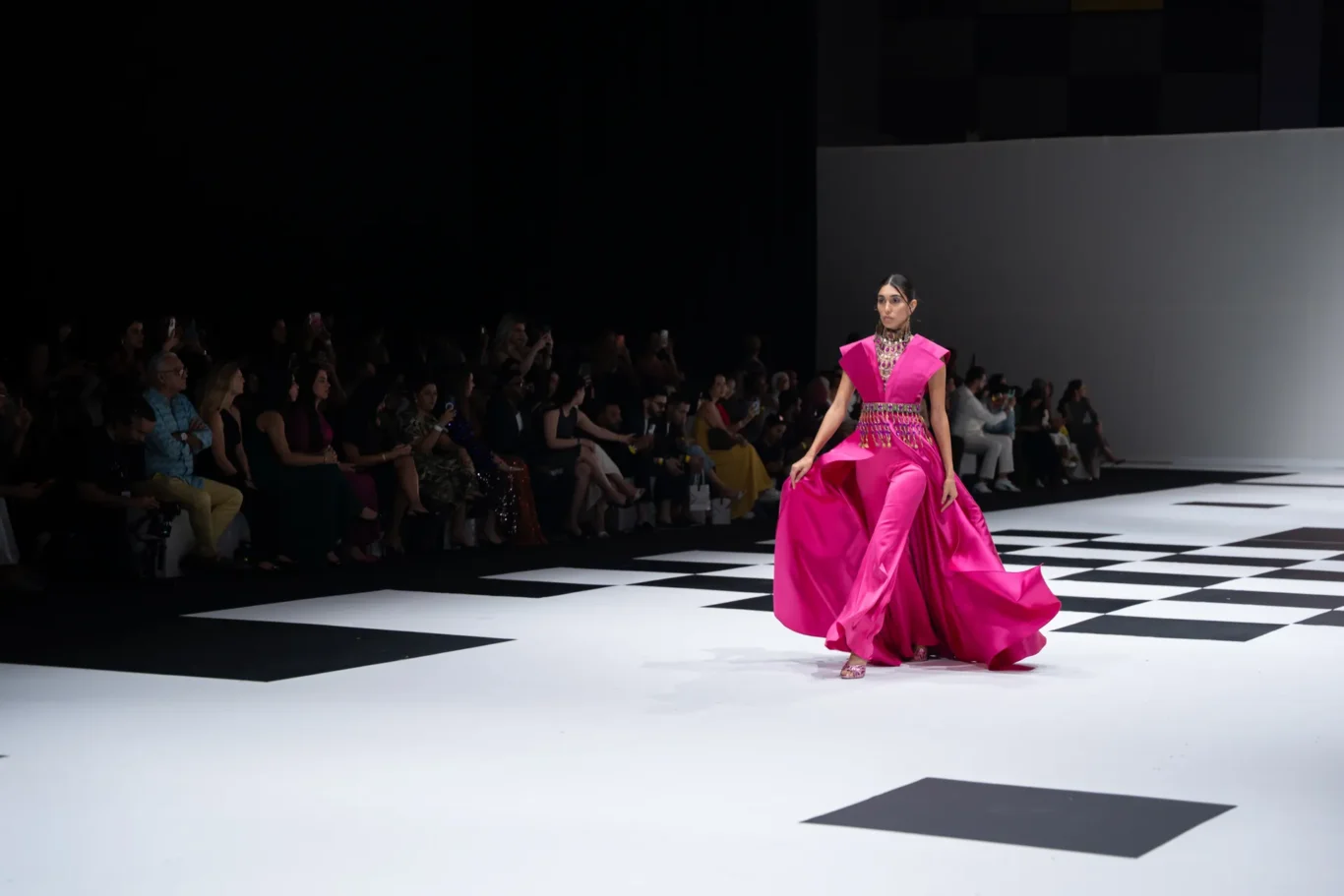 Model in a bright pink gown walks the checkerboard runway as the audience watches from the left side of the catwalk.