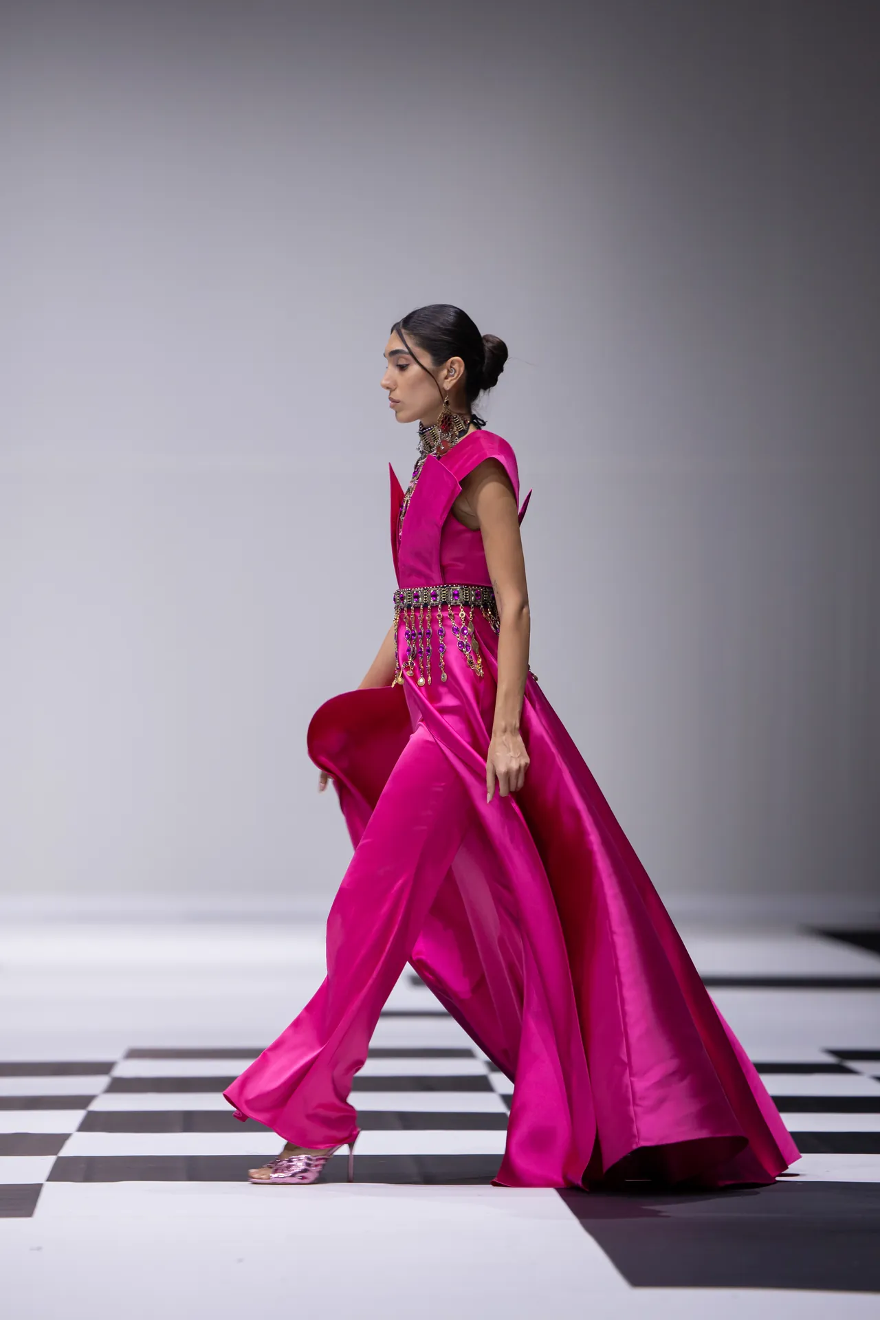 Female model in a vivid pink gown with a jeweled belt and choker walking the runway on a black-and-white checkered floor.