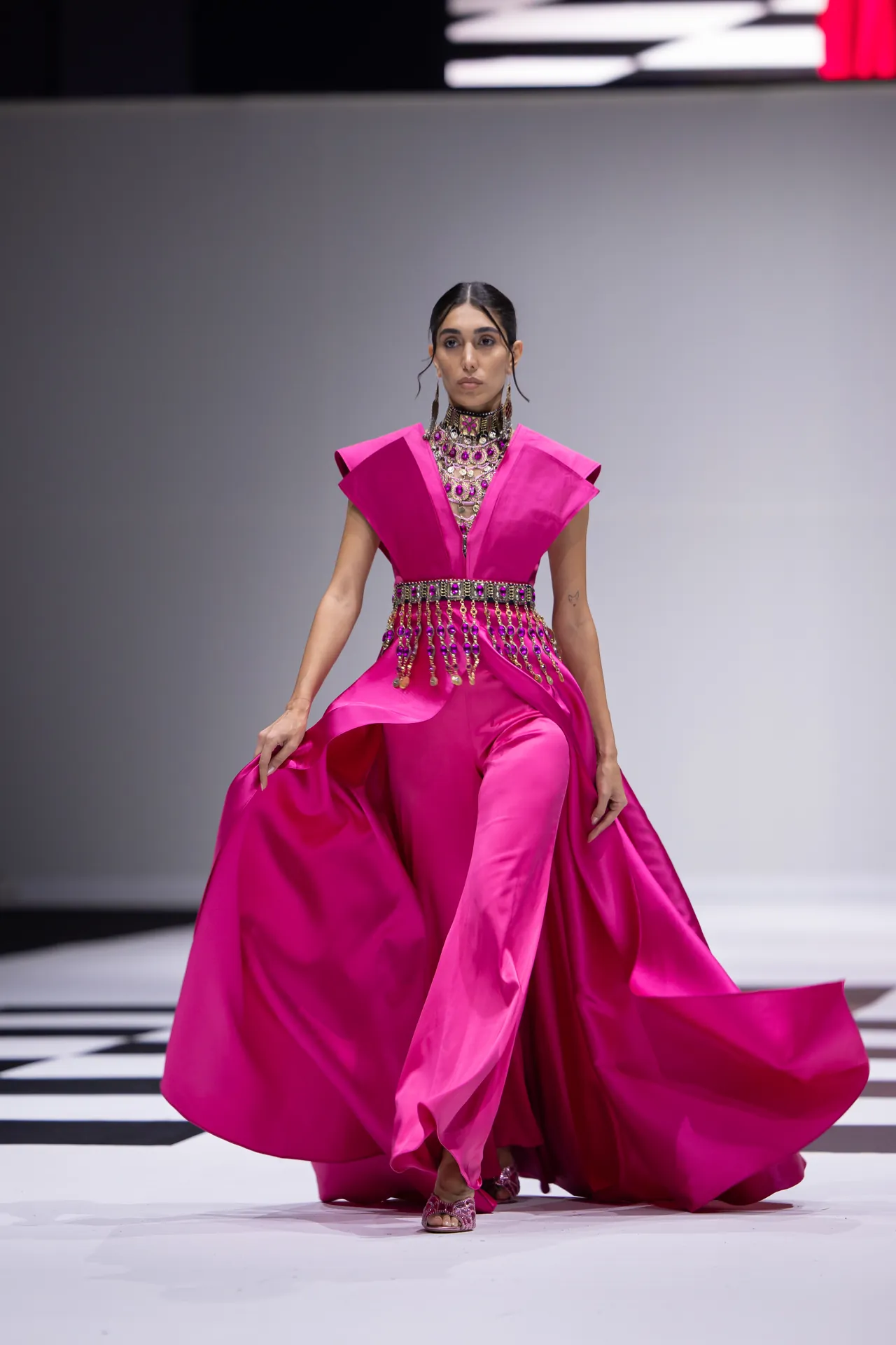Model struts a vivid pink gown with voluminous skirt and ornate jeweled belt on a runway, mid-walk stretch.