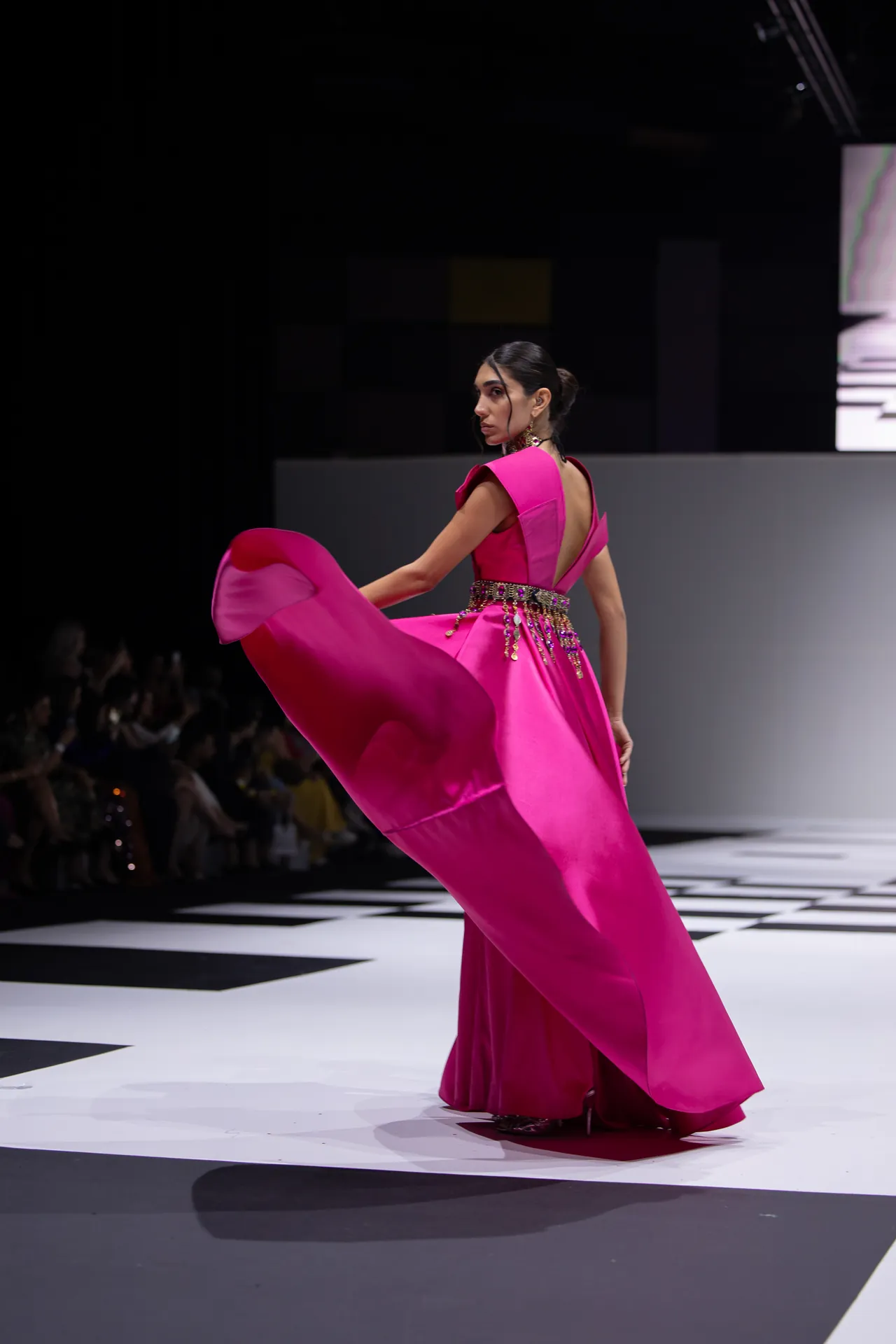 Model in vivid pink gown spins on a checkered runway during a fashion show, fabric flowing.