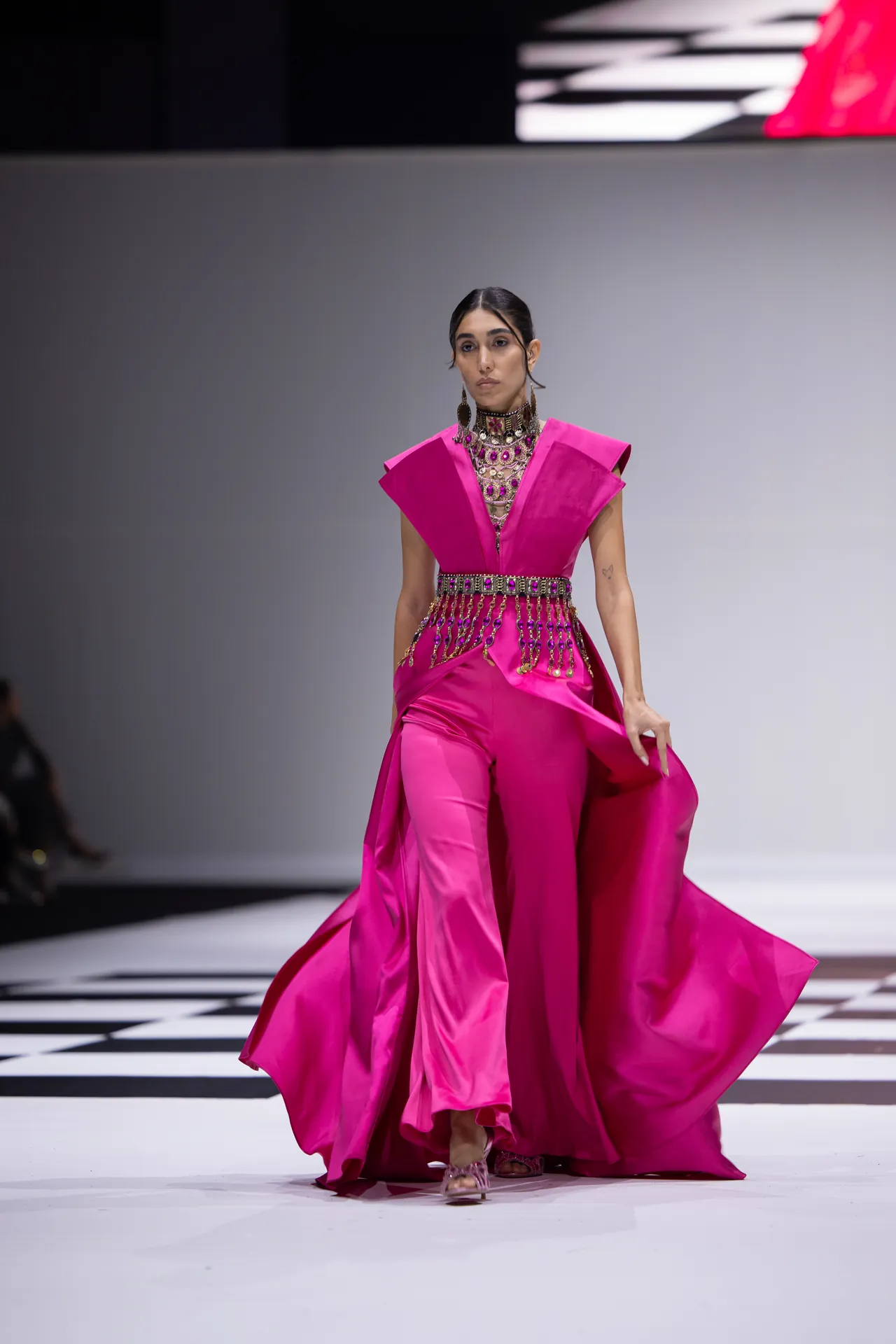 Model walks the runway in a vivid pink ensemble with cape-like shoulders and a jeweled belt, jewelry adorning the neck and chest area.