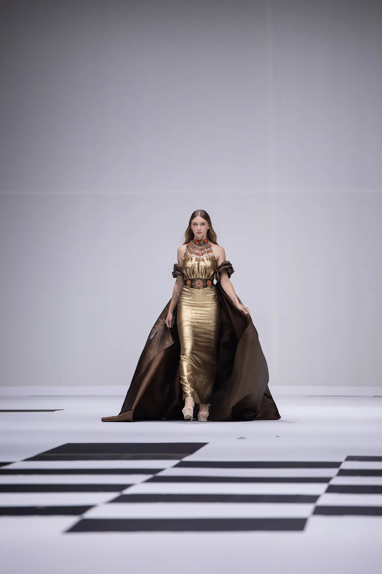 Model on a runway wearing a gold metallic gown with a voluminous brown cape and ornate jewelry.
