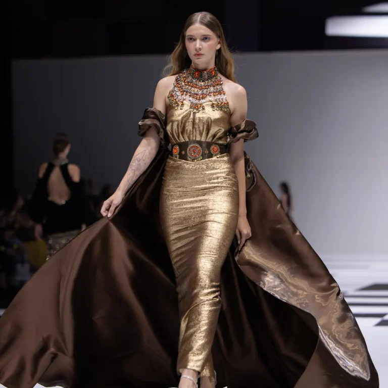 Model walks a runway in a gold metallic gown with an ornate beaded collar and a flowing brown cape behind her.