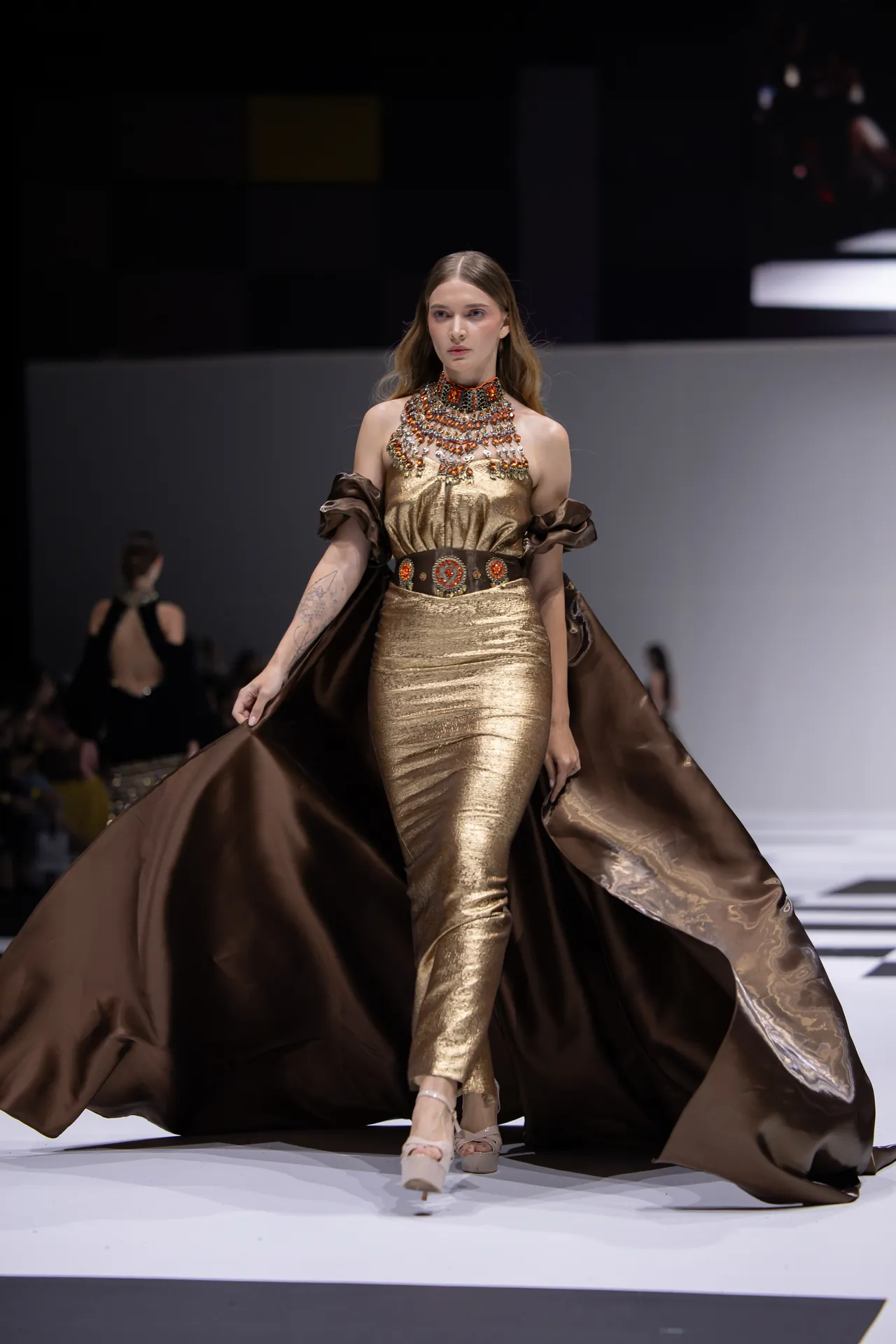 Model walks a runway in a gold metallic gown with an ornate beaded collar and a flowing brown cape behind her.