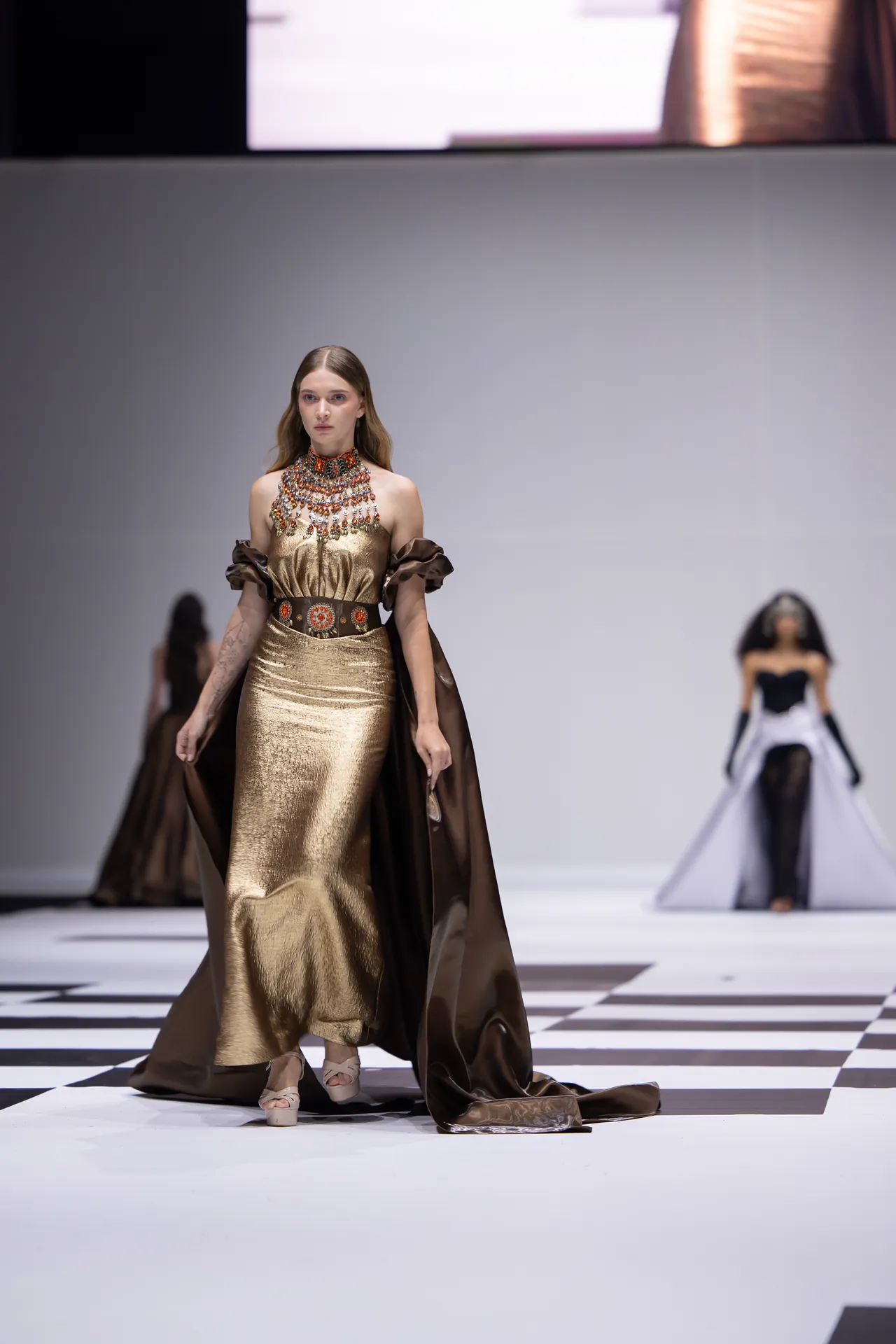 Model wearing a gold metallic gown with a jeweled collar and brown cape walking a black-and-white checkered runway at a fashion show