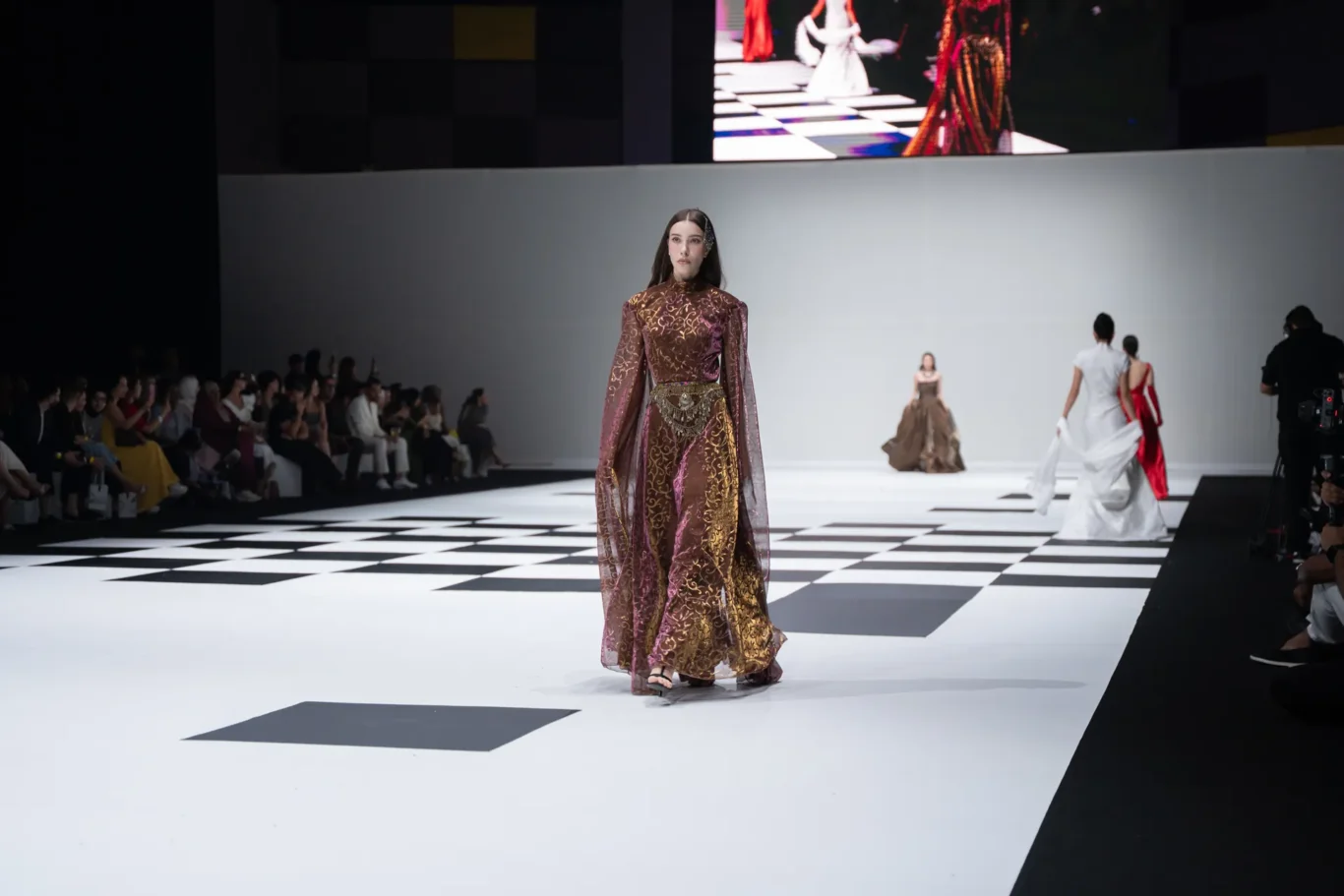Model walks down a black-and-white checkered runway in an ornate brown and gold gown with a jeweled belt, audience on the left watching.