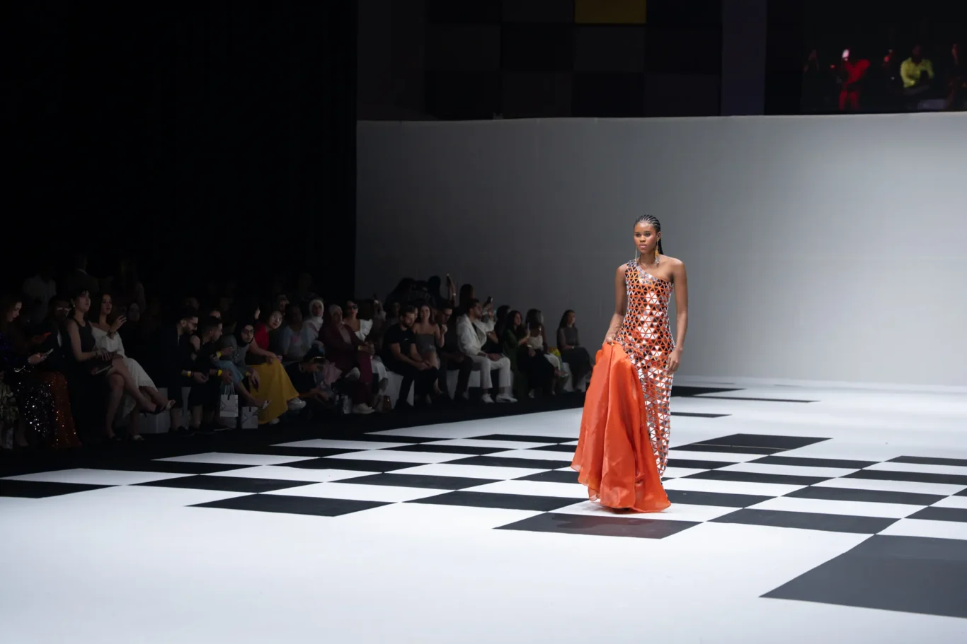 Model walks the runway in an orange geometric dress, holding a matching orange cape; audience sits along the catwalk.